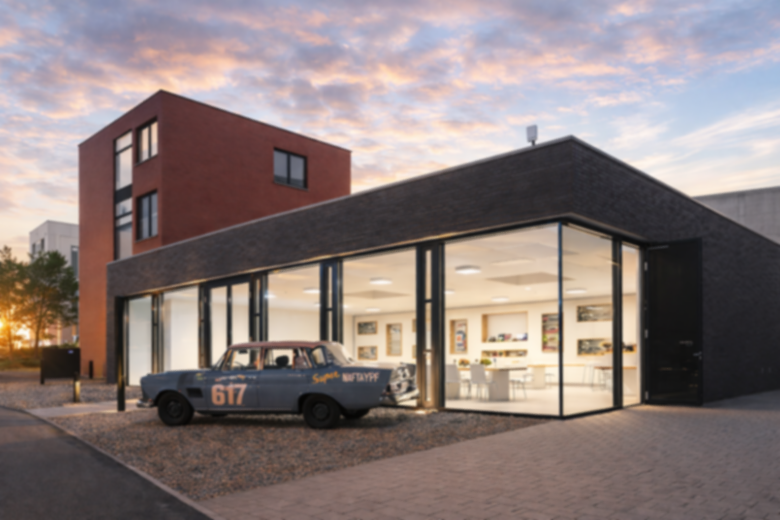 Modernes Geschäftshaus mit großem Fenster, in dem Bücherregale sichtbar sind, und einem alten, beschrifteten Auto davor, bei Sonnenuntergang.