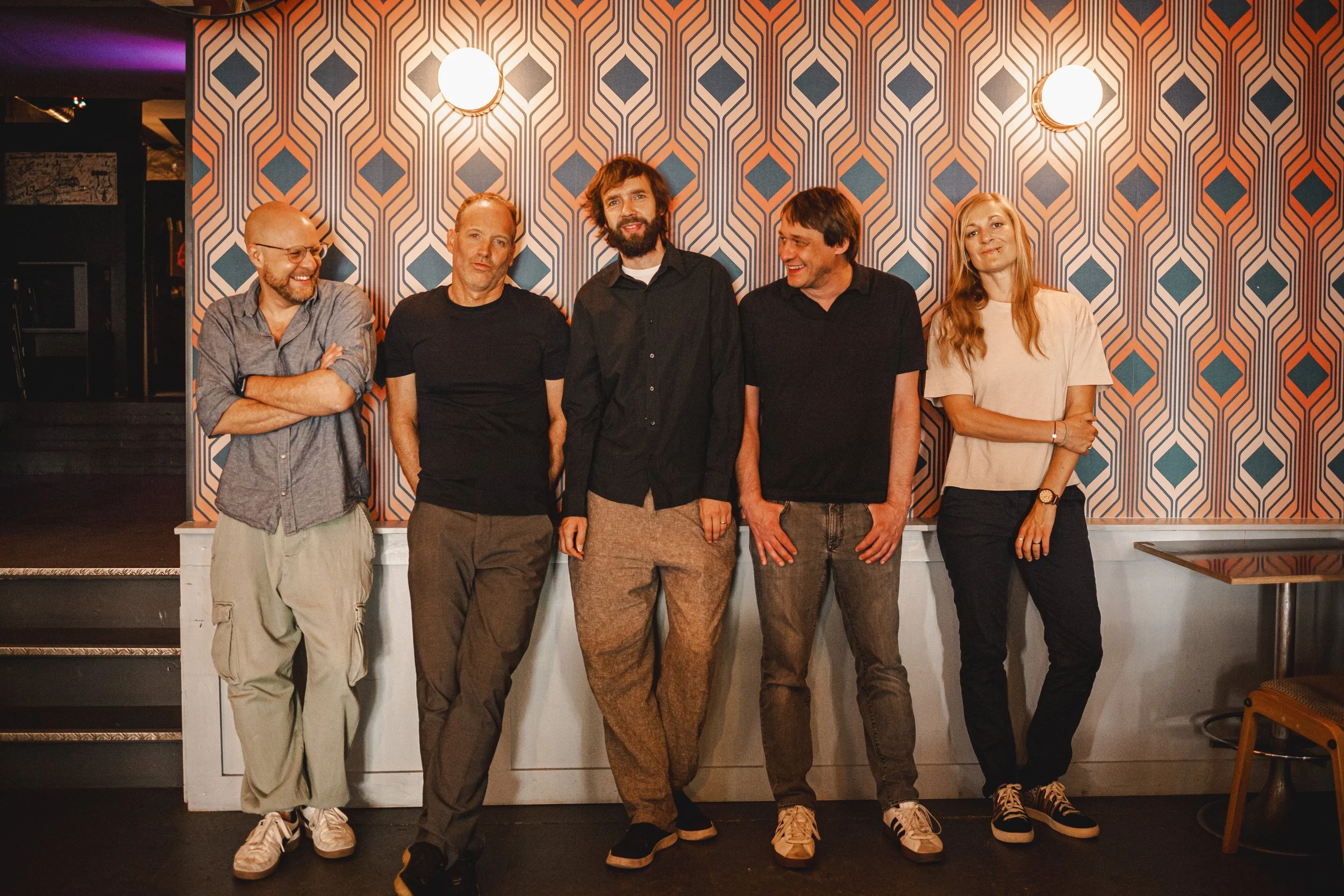 Eine Gruppe von fünf Personen steht vor einer bunten Tapete und lächelt in die Kamera