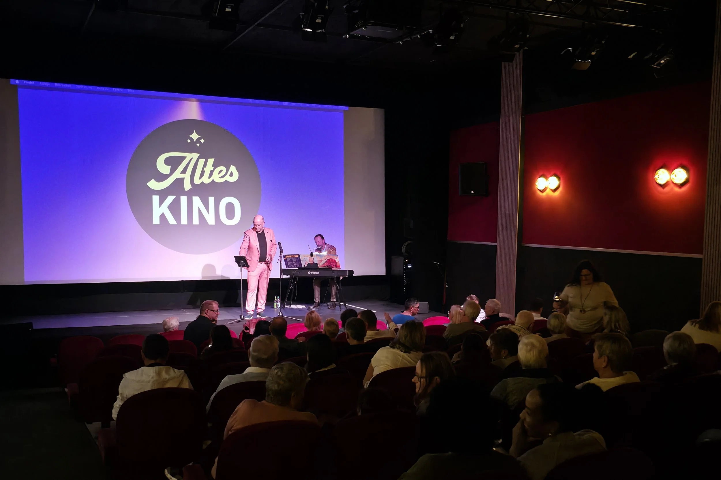 Ein Musiker steht auf einer Bühne vor einer Leinwand auf der Altes Kino zu lesen ist