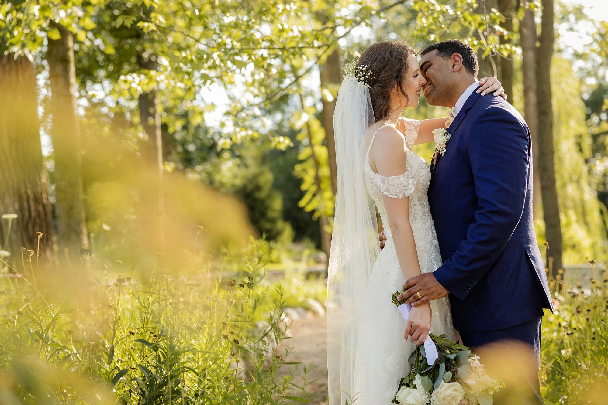 Bride and groom embracing in a garden setting, wearing wedding attire, with lush greenery and sunlight filtering through trees.