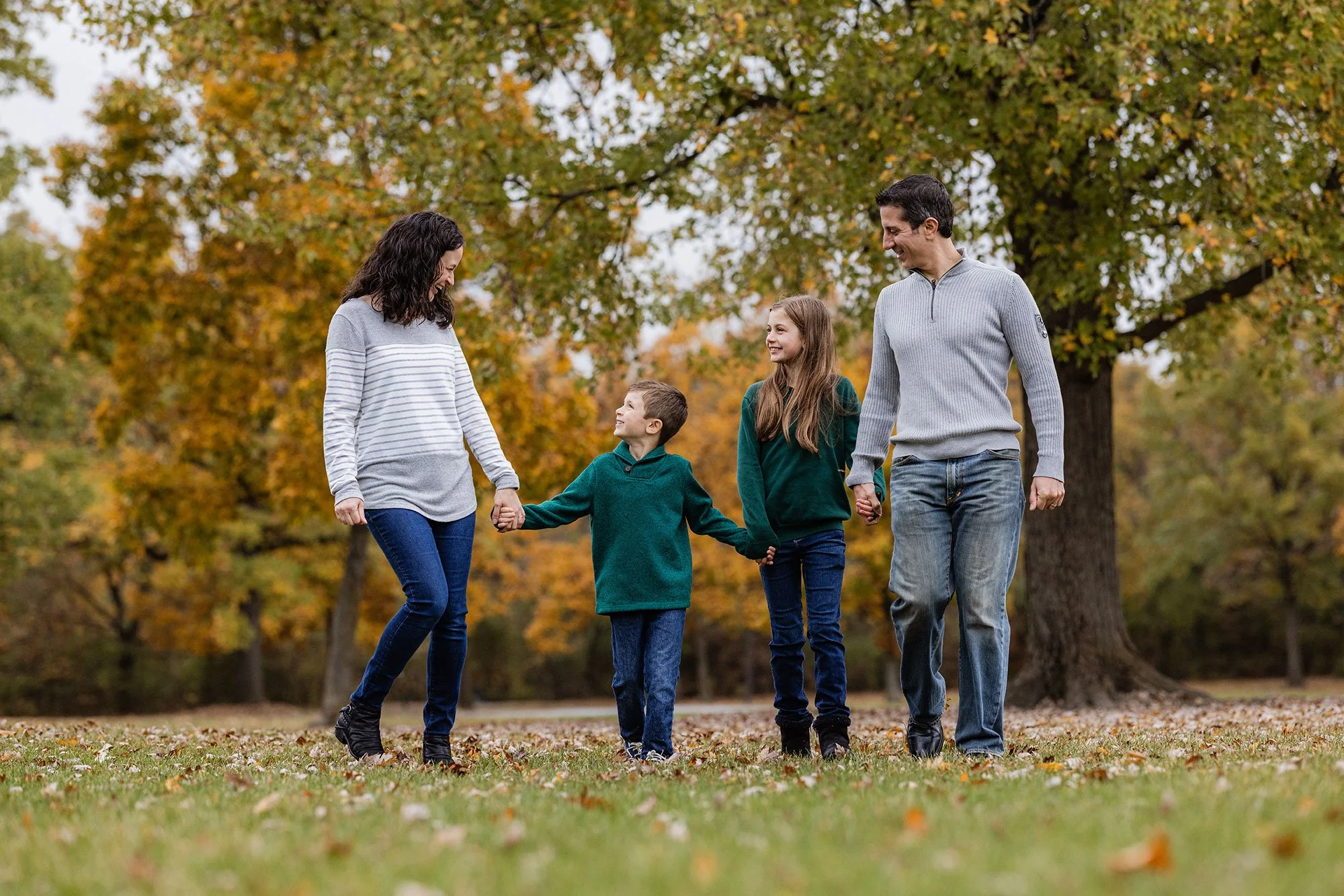A family of four, consisting of two adults and two children, walking hand in hand through a park with green grass and autumn trees in the background.