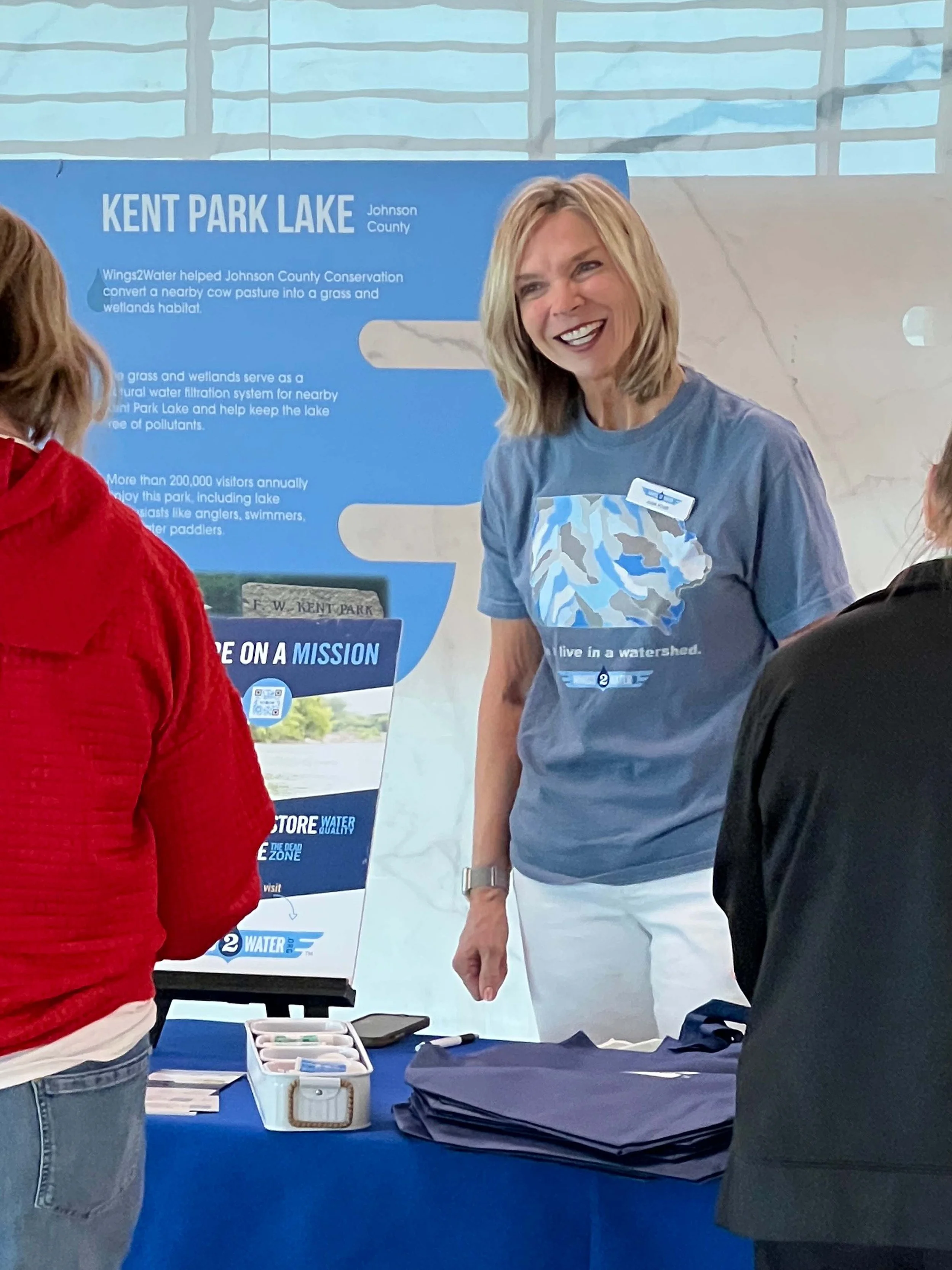 A woman with blonde hair wearing a blue t-shirt with a bear graphic and a name tag smiles and talks to two people at an informational booth about Kent Park Lake in Johnson County. There are blue display panels, a table with brochures, and folders.