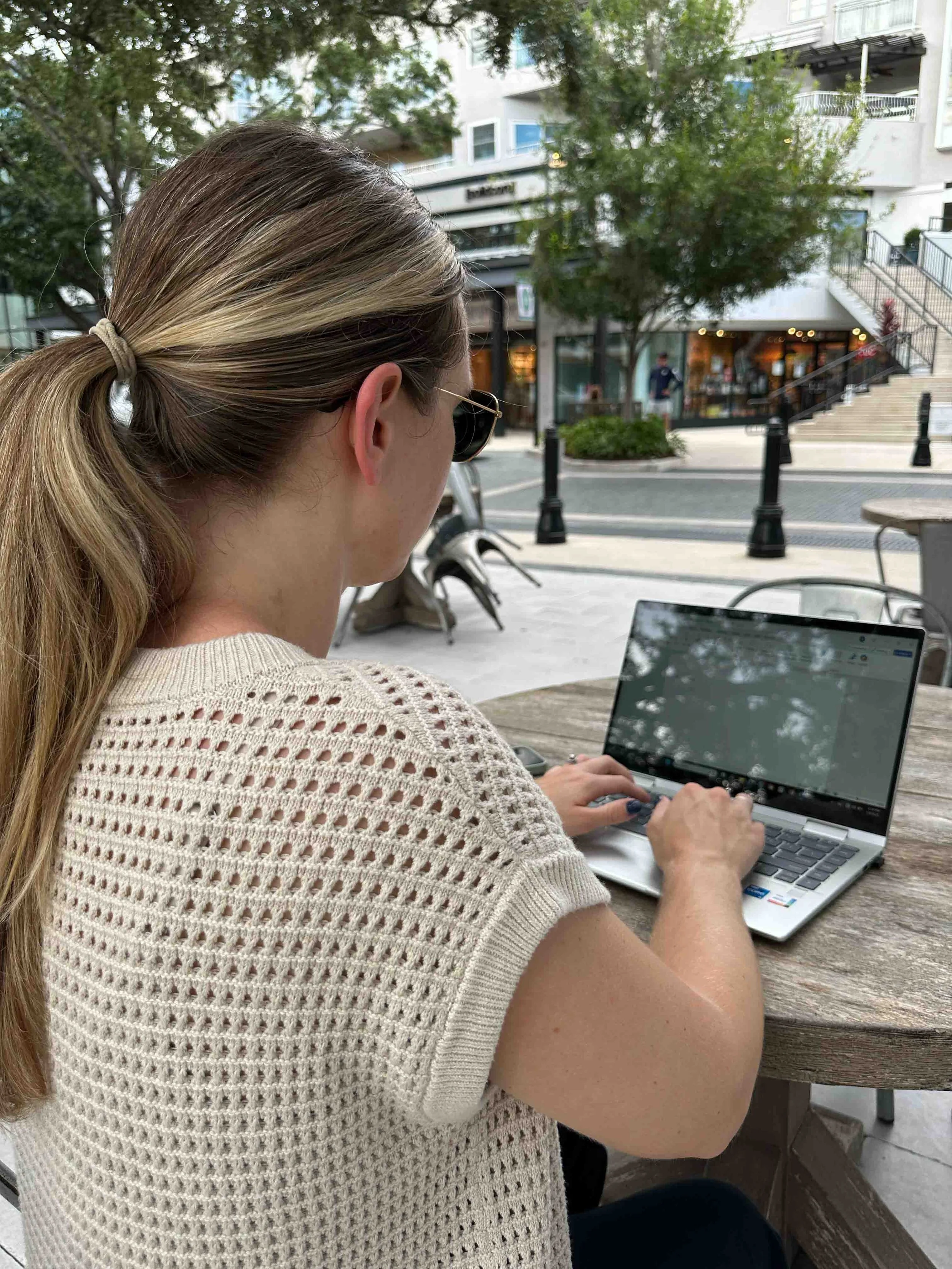 A woman with long blonde hair tied in a ponytail, wearing sunglasses and a cream-colored knit top, sitting outdoors at a wooden table, working on a silver laptop, with trees, a building, and people in the background.