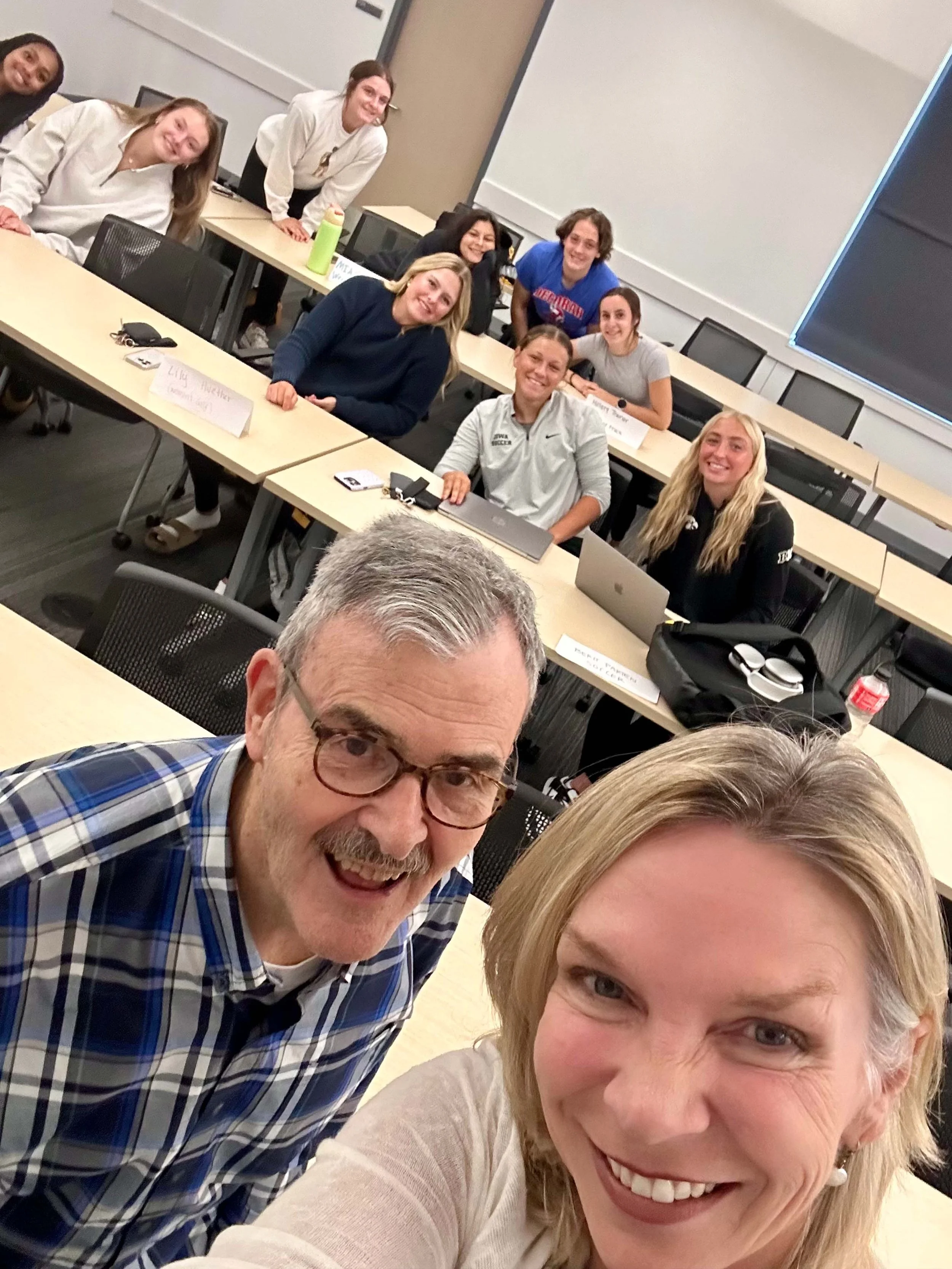 Smiling group of women and a man taking a selfie in a classroom or conference room. The background shows more people sitting at tables, some with laptops and notebooks.