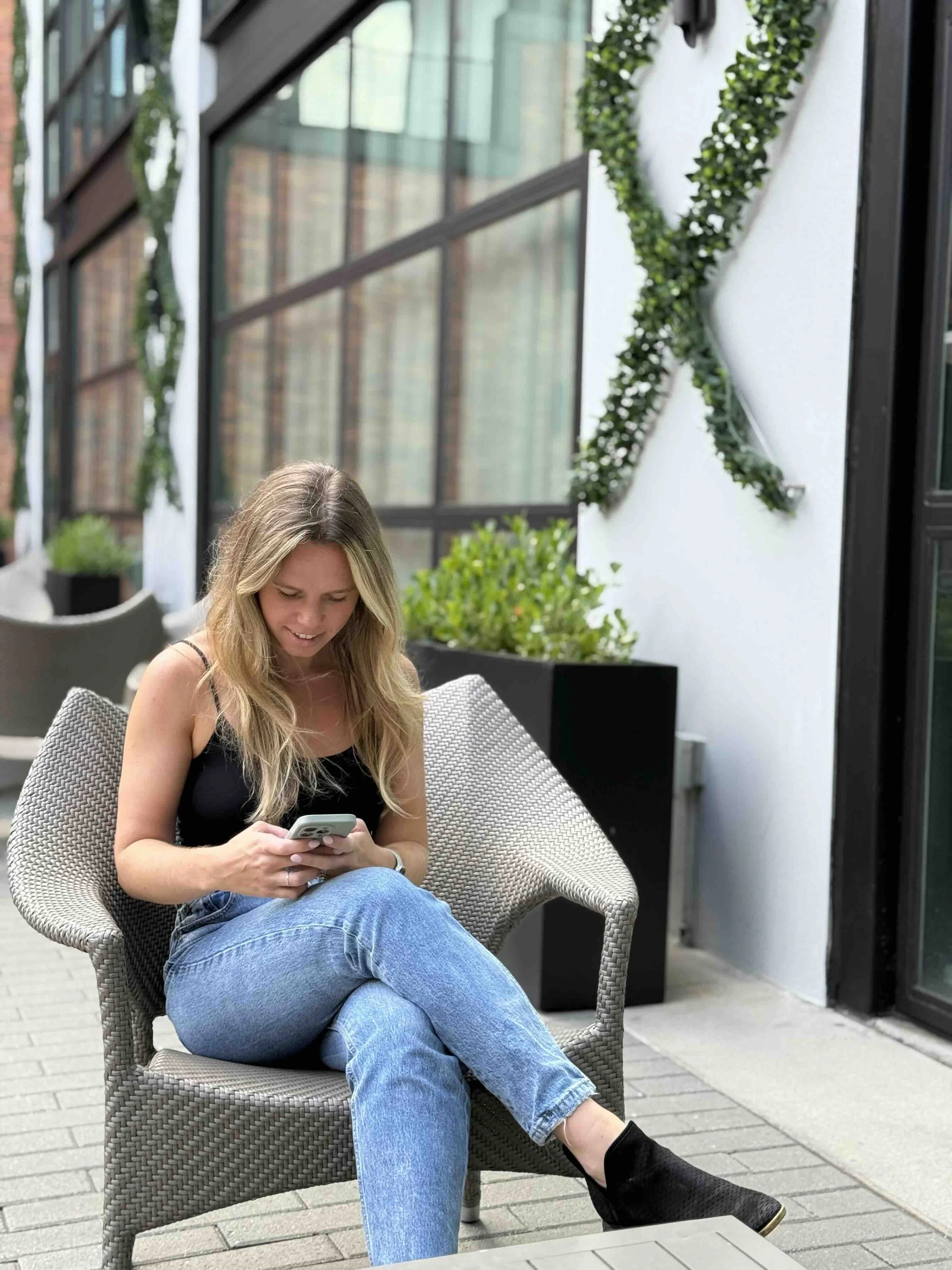 A young woman with blonde hair, wearing a black top and blue jeans, is sitting on a woven outdoor chair while looking at her smartphone. She has her legs crossed with one foot in a black boot, on a patio area with potted plants and a modern building with glass windows in the background.