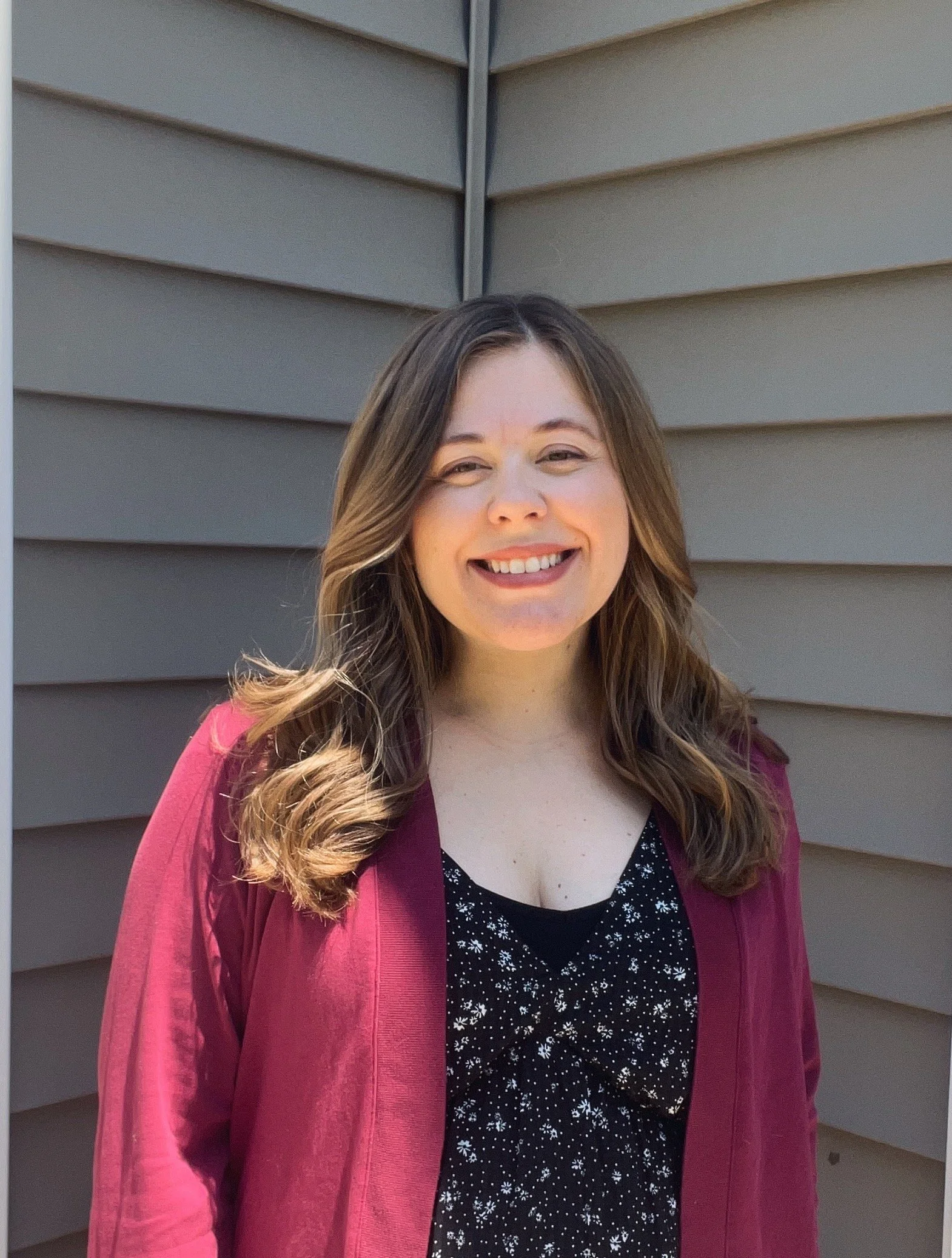 A young woman with long wavy brown hair, smiling, standing outdoors in front of beige horizontal siding. Speech therapy, speech therapist, pediatric speech therapy, speech language pathologist.