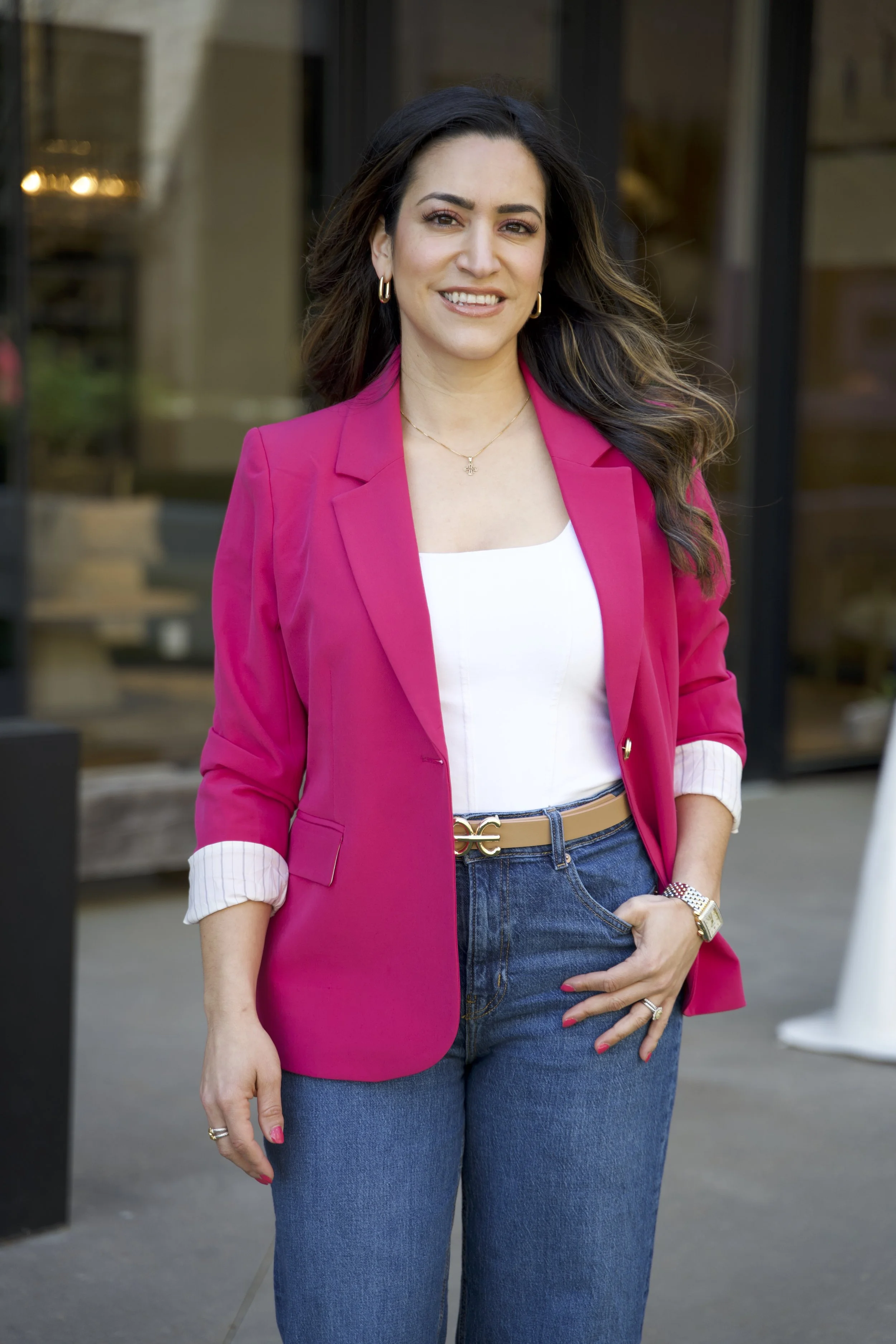 A woman with long dark hair wearing a bright pink blazer over a white top and blue jeans, standing outdoors in front of a glass building, smiling at the camera. Speech therapy, pediatric speech therapy, speech language pathologist.