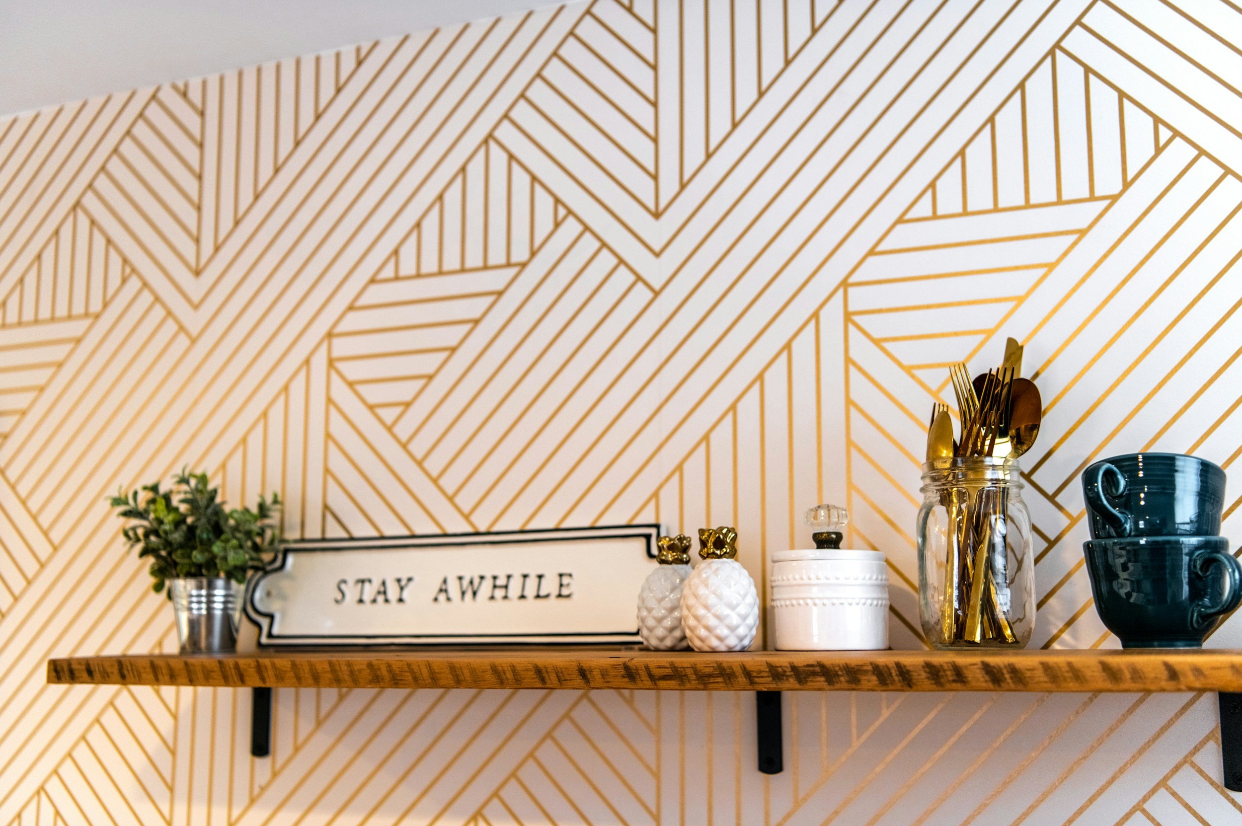 Decorative wooden shelf with a sign reading 'STAY AWHILE', a potted plant, small pineapple-shaped decor, a white container, a jar filled with gold utensils, and blue cups against a geometric patterned wall.