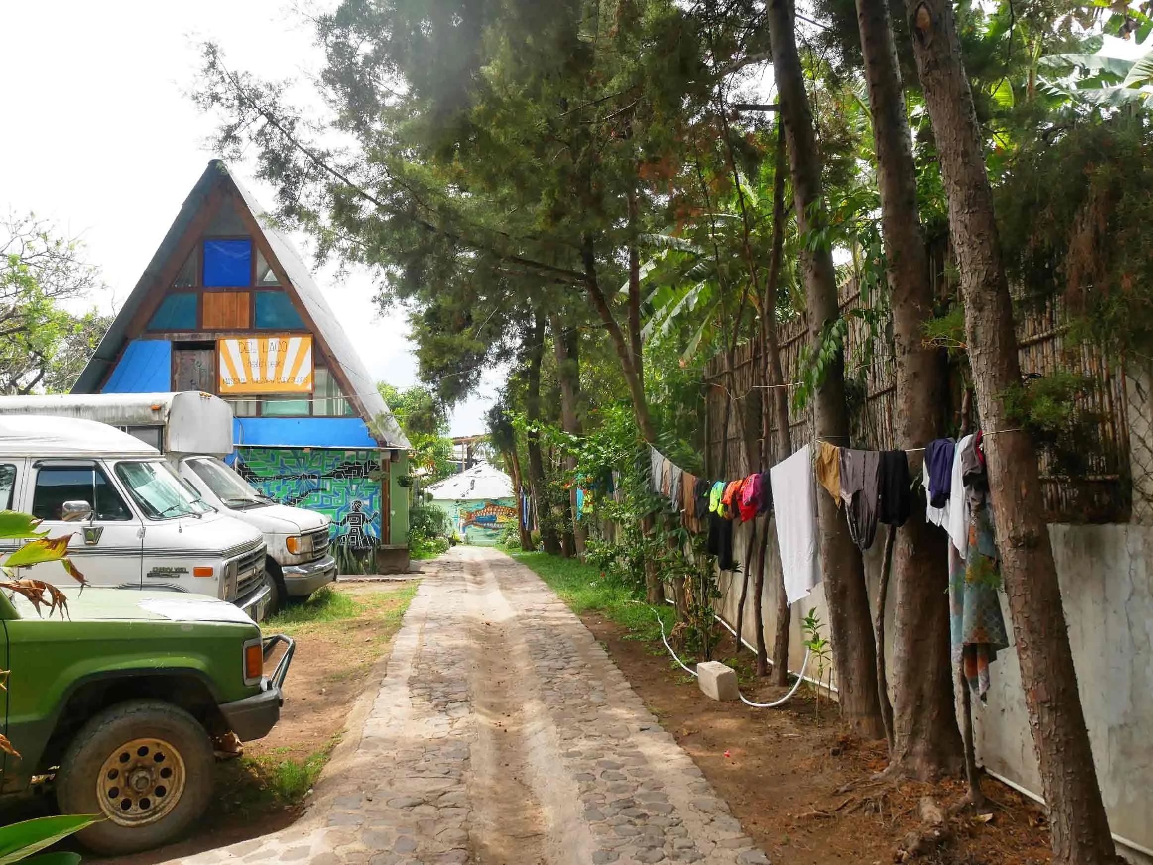 A narrow cobblestone alleyway with tall trees on the right and parked cars on the left. A colorful triangular building with murals and a sign that reads 'DEL LAGO' is at the end of the alley. Clothes are hanging on a line tied between trees on the ri