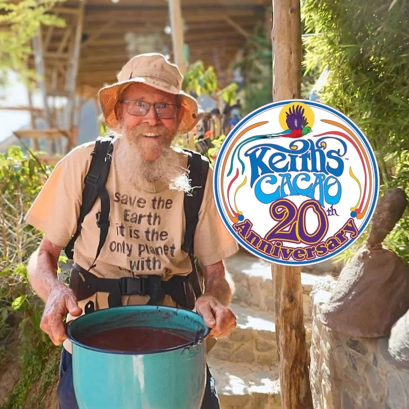 An elderly man with a long white beard, hat, glasses, and backpack holding a large blue container of cacao, standing outdoors near trees and a stone pathway, celebrating Keim's Cacao 20th anniversary