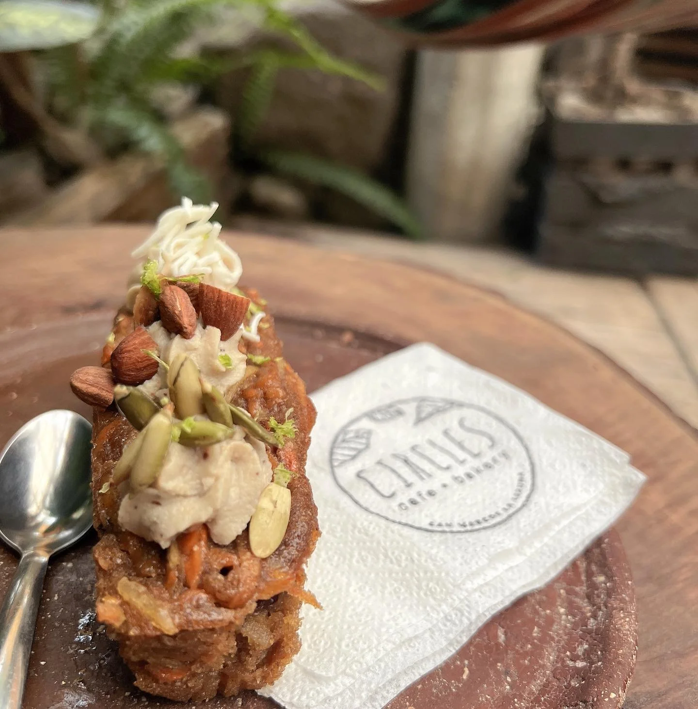 A dessert topped with assorted nuts and sesame seeds, served on a wooden plate with a silver spoon and a napkin that has a logo, in a rustic setting.