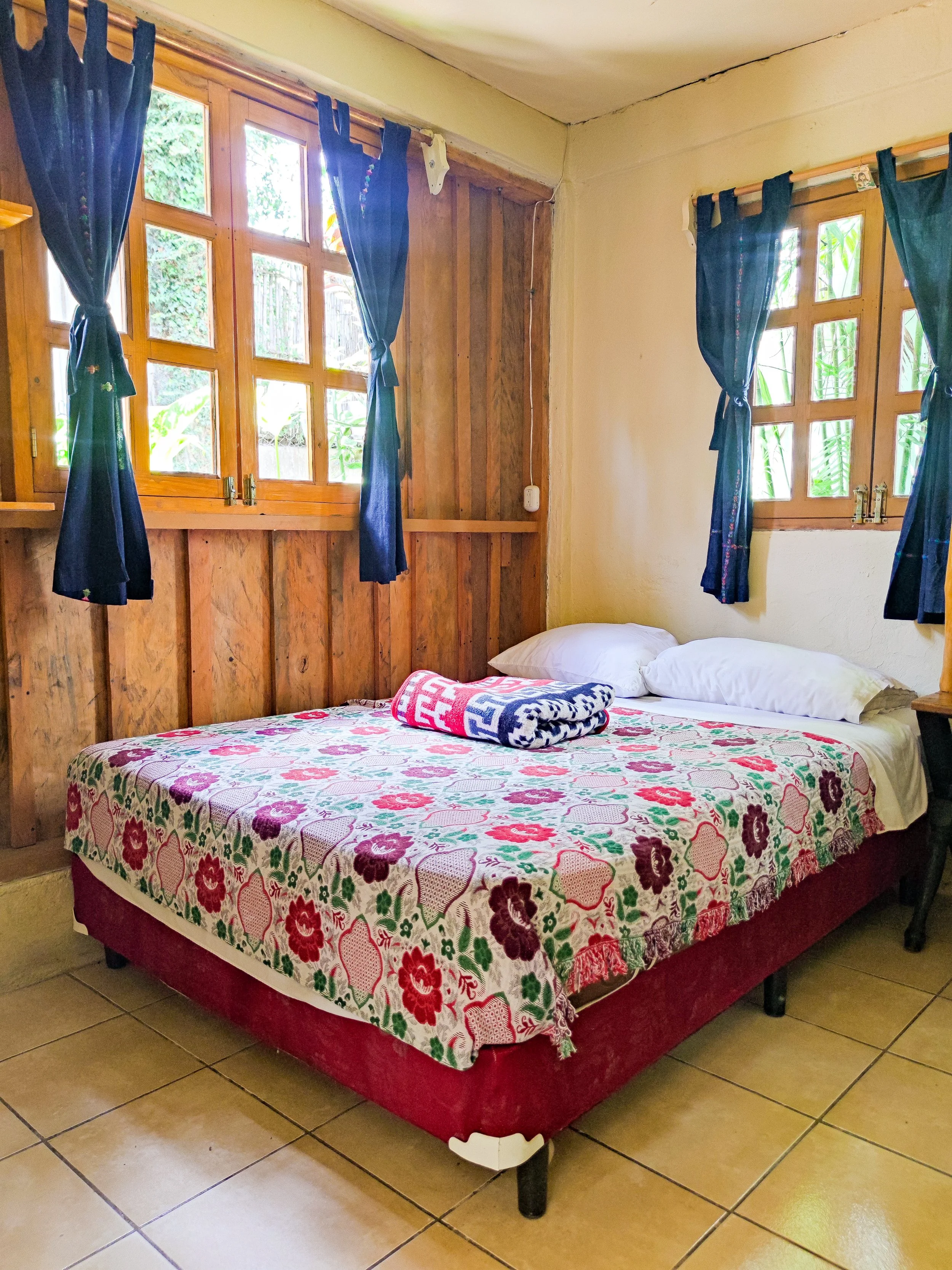 A bedroom with a bed covered by a floral bedspread, two pillows, a rolled towel, and surrounded by wooden window frames with blue curtains.