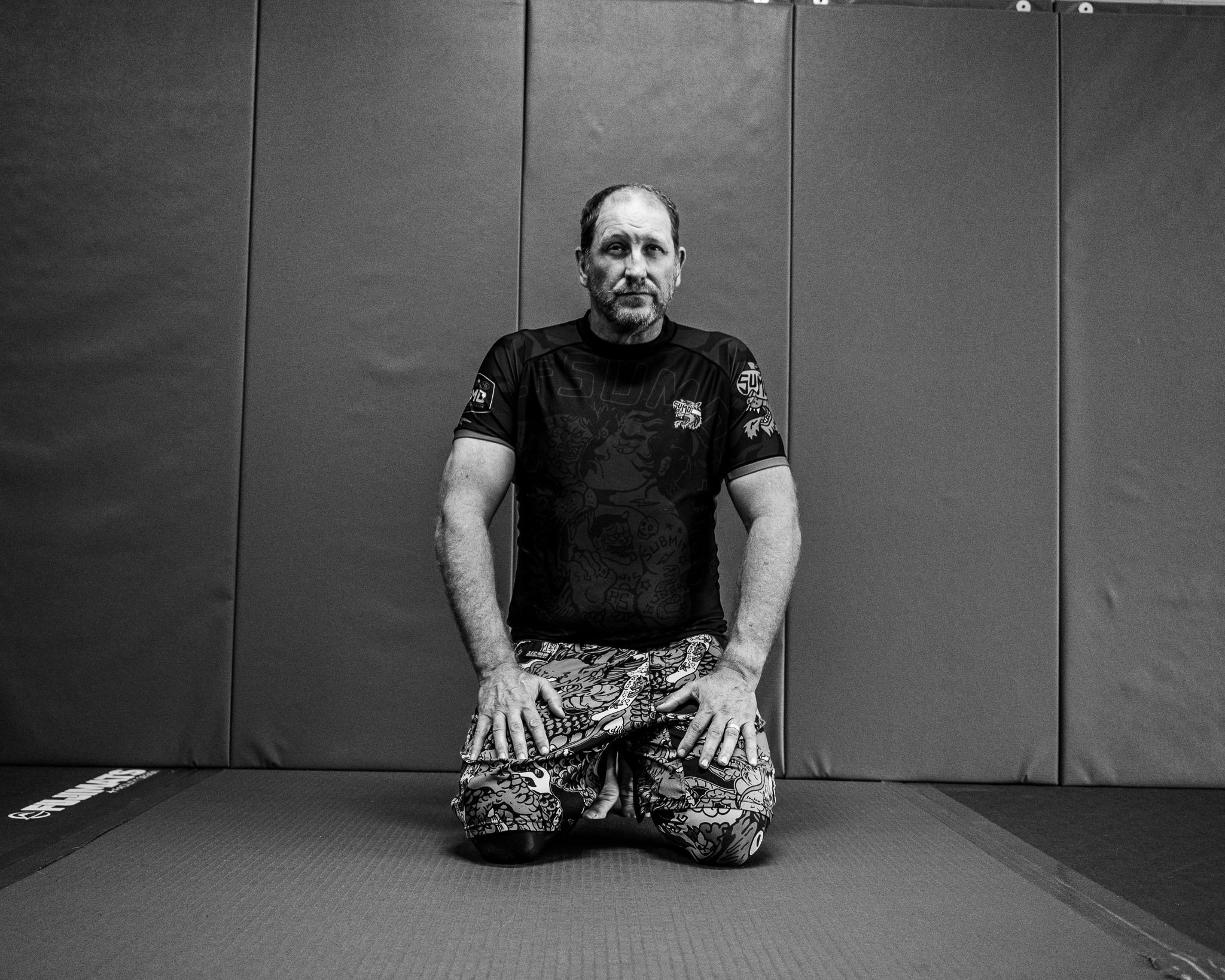 A man with a beard and short hair sitting cross-legged in a martial arts gym, wearing a martial arts uniform with various patches and design. He is in a kneeling position on a black mat, in front of a gray padded wall.