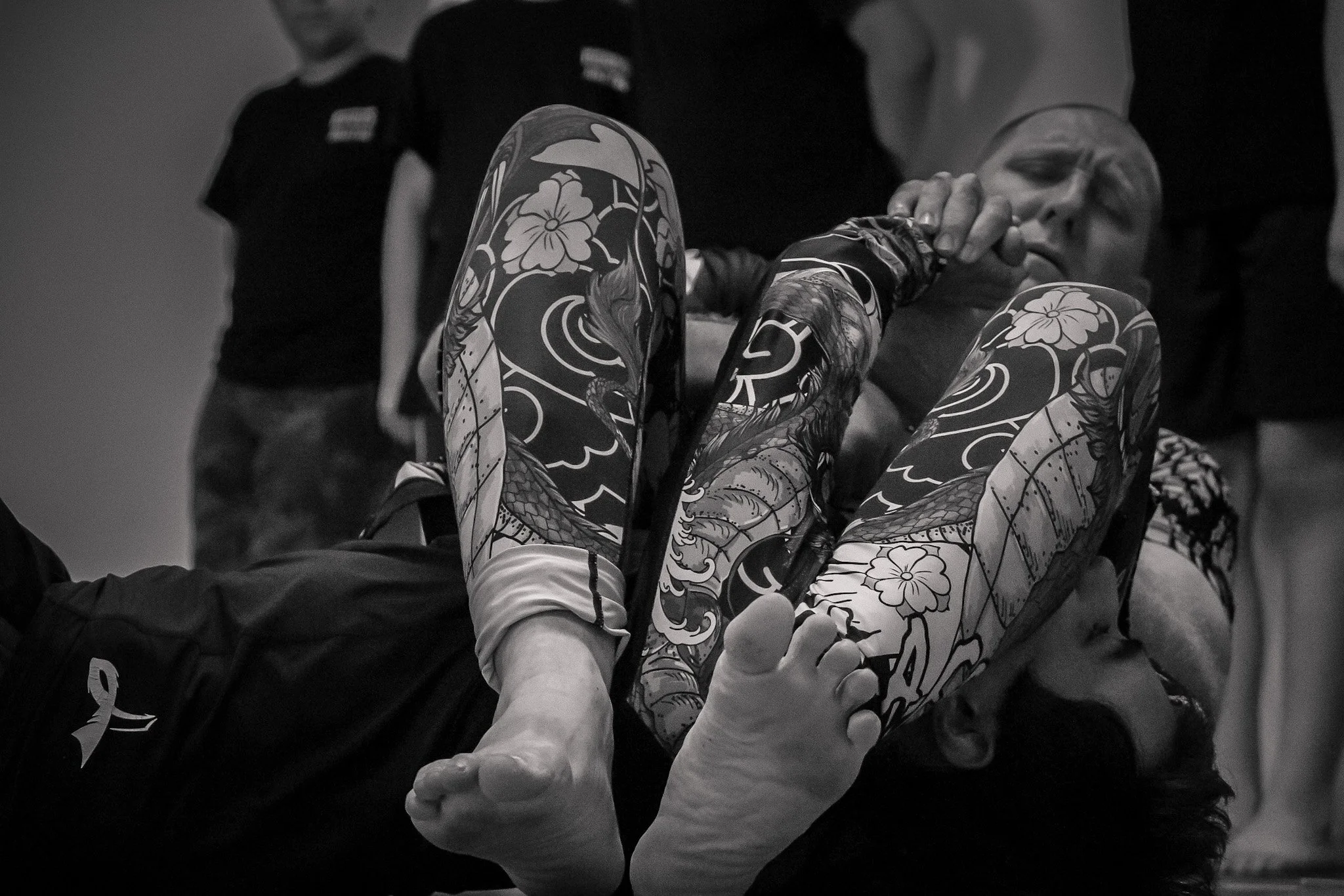 A woman demonstrating Brazilian Jiu-Jitsu techniques with a tattooed leg, while a man applies a headlock. Several people are observing in the background.