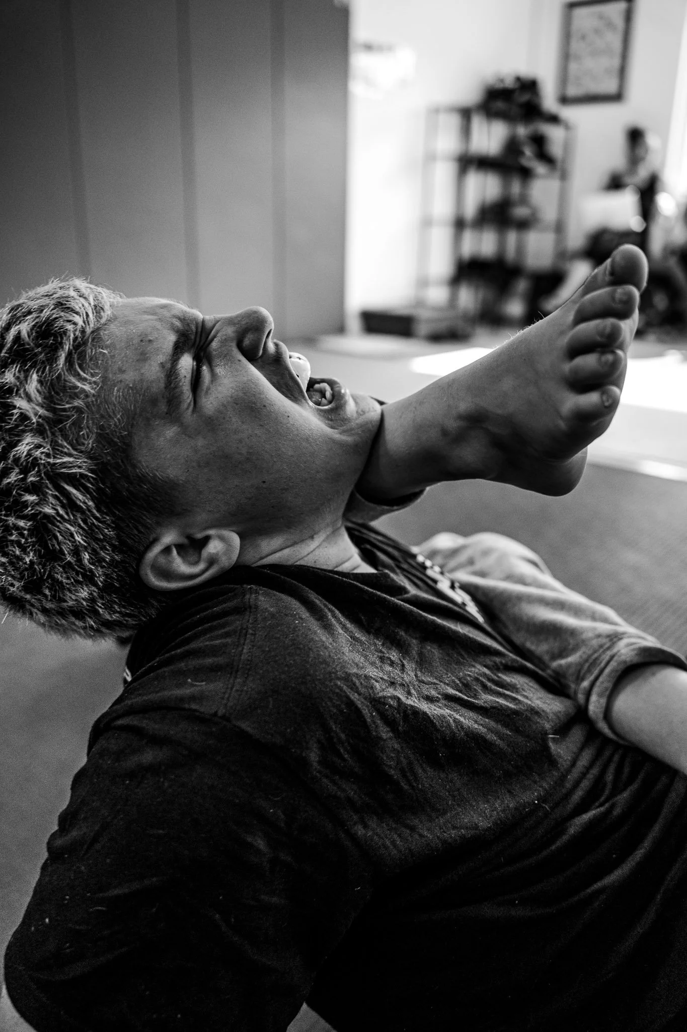 A young person with curly hair is laughing or smiling, lying down or reclining, with their head turned to the side and mouth open. The background shows a room with a shelving unit and a person sitting in the distance.