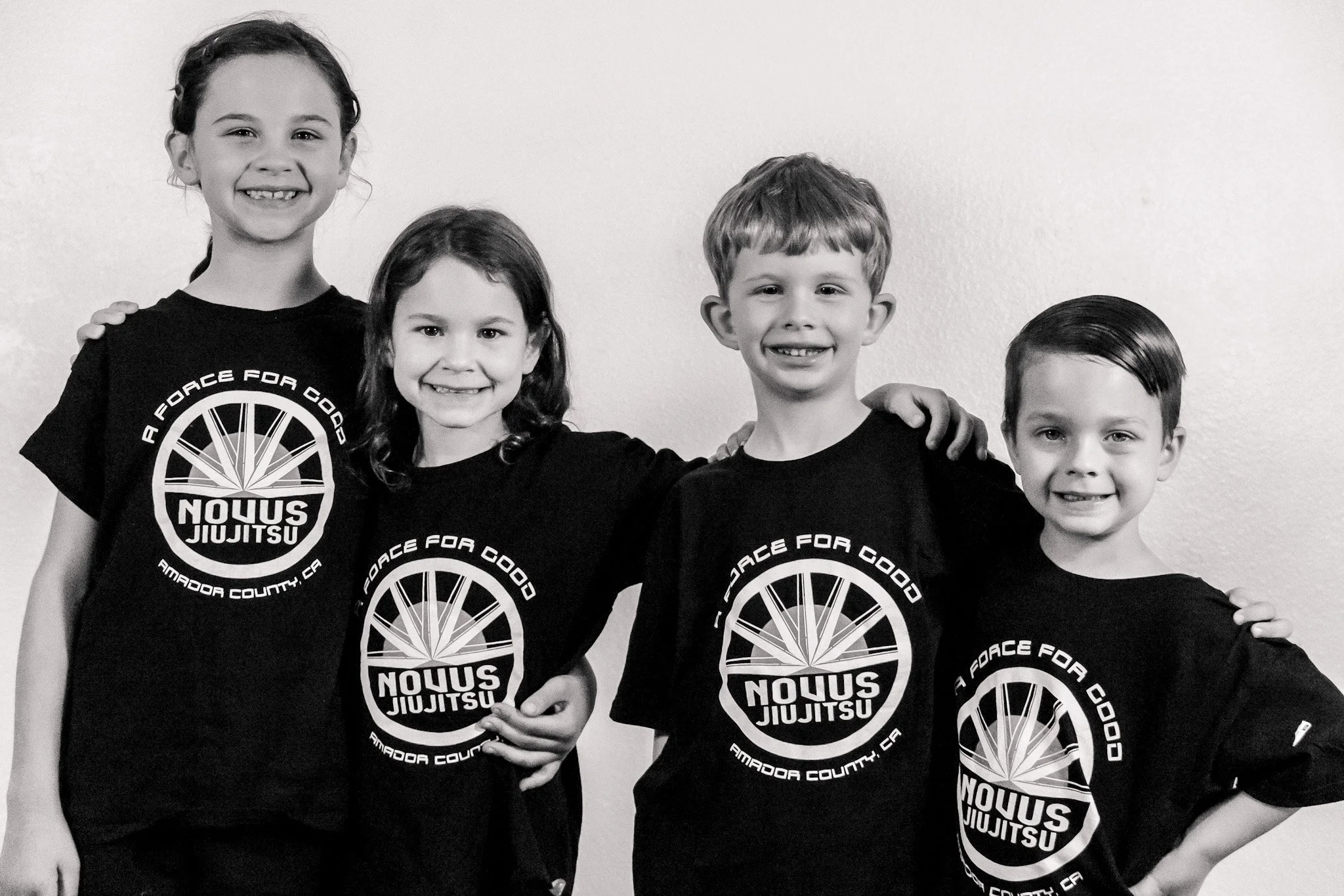 Four children standing side by side, smiling, wearing matching black t-shirts with a logo that reads "A Force for Good Isso Jujitsu Amador County, CA". They have their arms around each other's shoulders against a plain background.