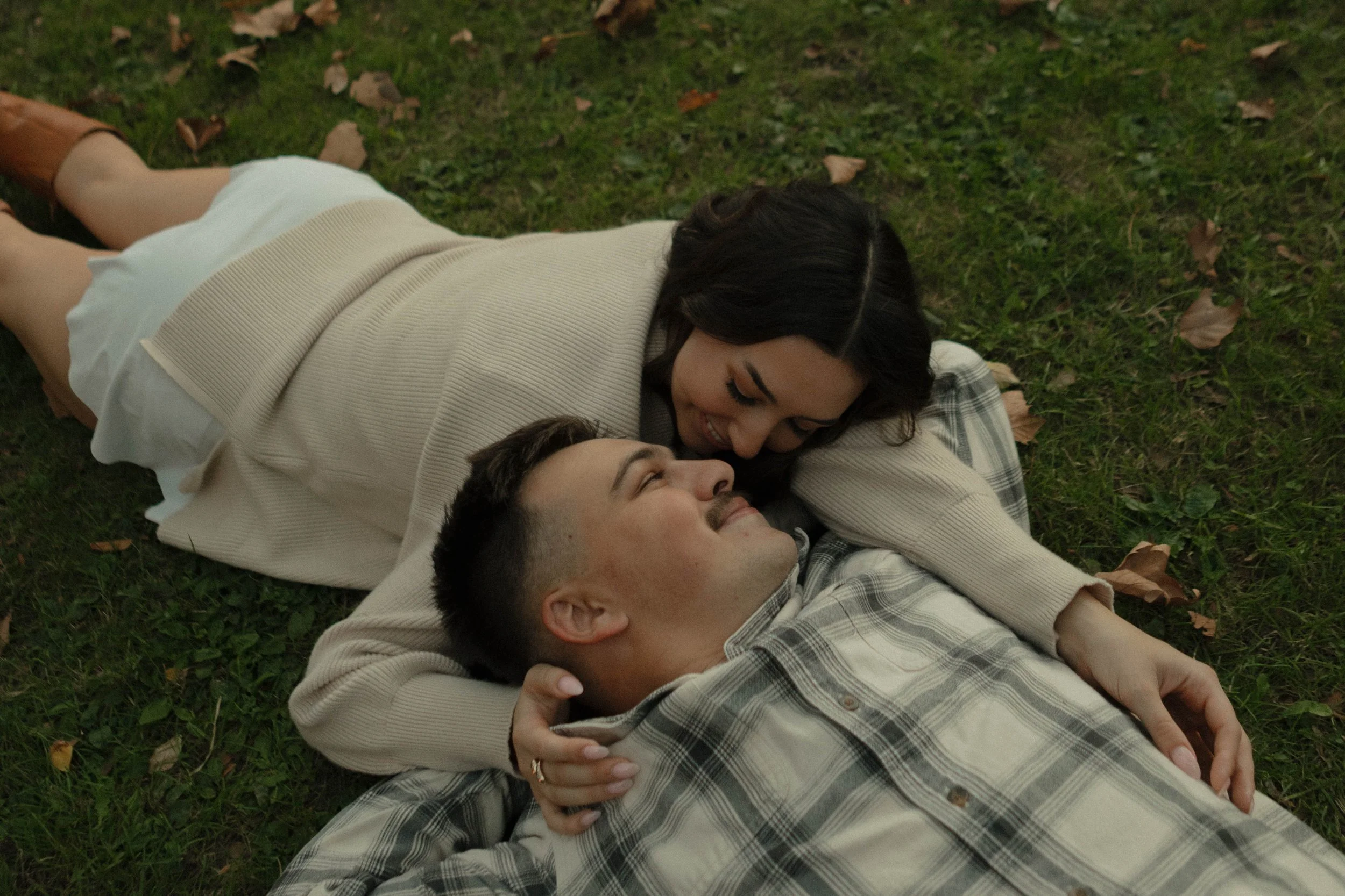A young woman and a young man lying on grass, smiling and looking at each other, with autumn leaves scattered around.