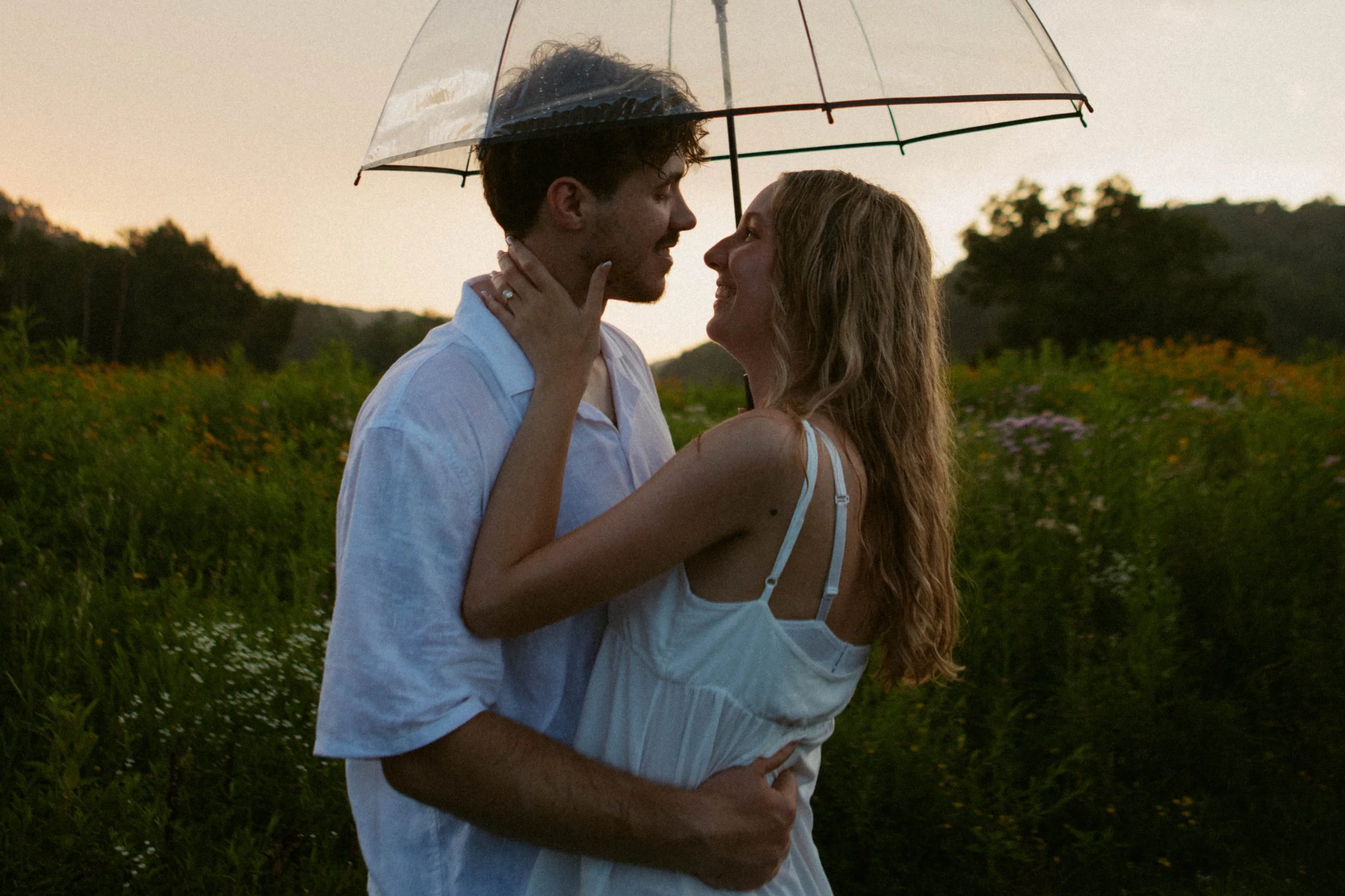 A young couple standing close together outdoors under a transparent umbrella, smiling and looking into each other's eyes during sunset in a field of flowers.