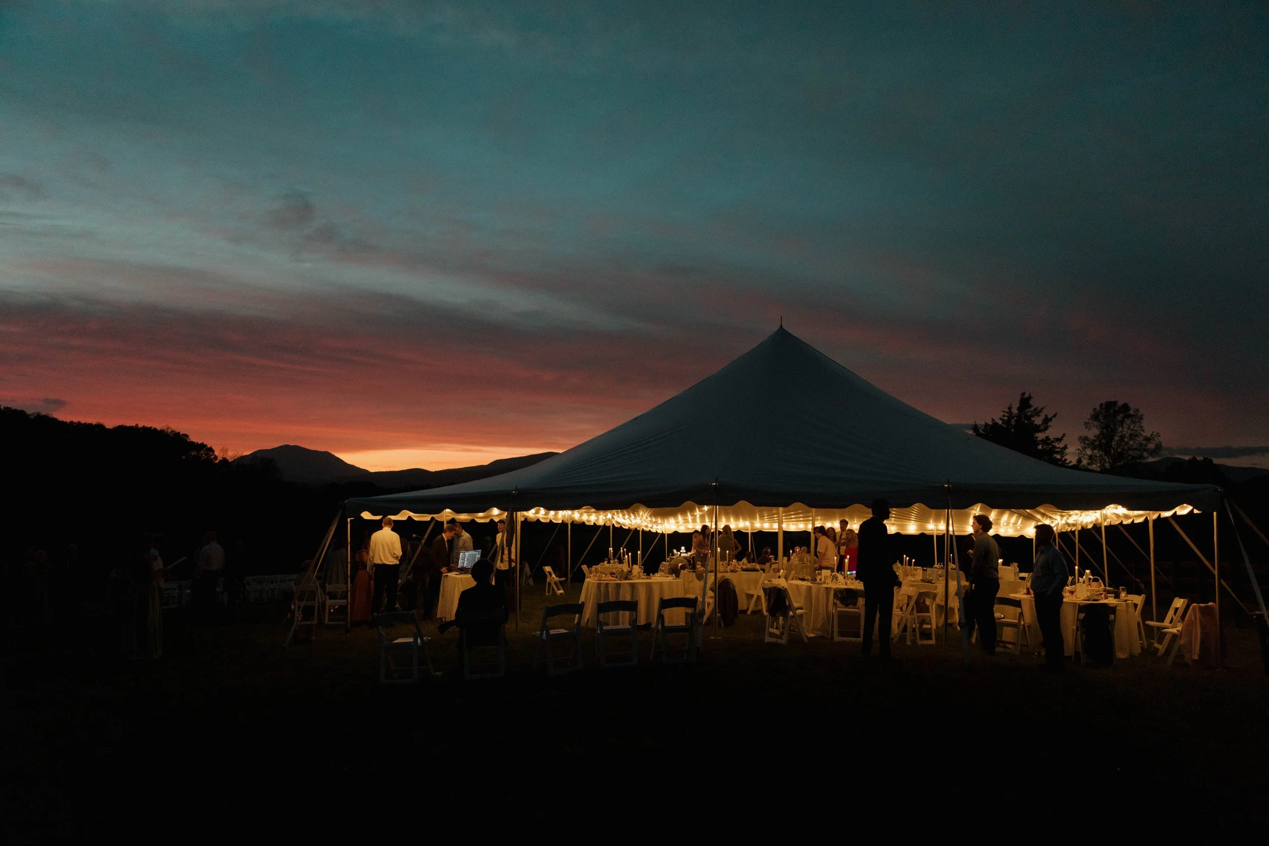 A large outdoor party tent illuminated with string lights at sunset, with people standing and sitting around tables set for an event.