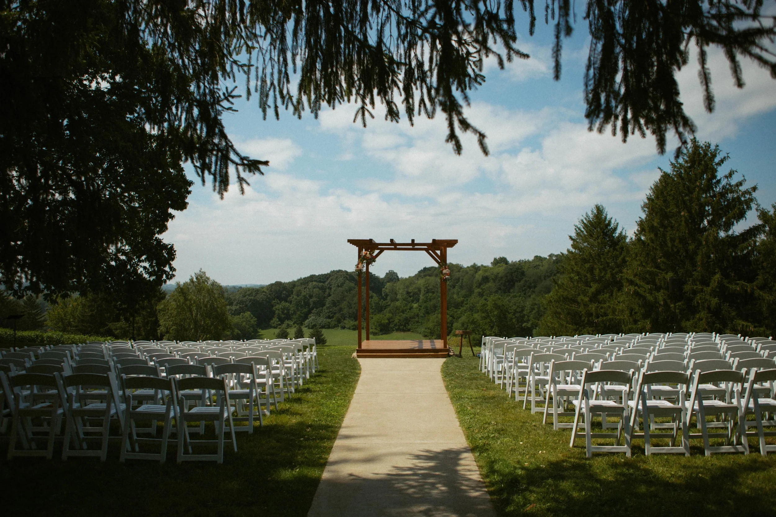 An outdoor wedding setup with rows of white chairs facing a wooden arch on a grassy field under a partly cloudy sky, surrounded by trees and greenery.