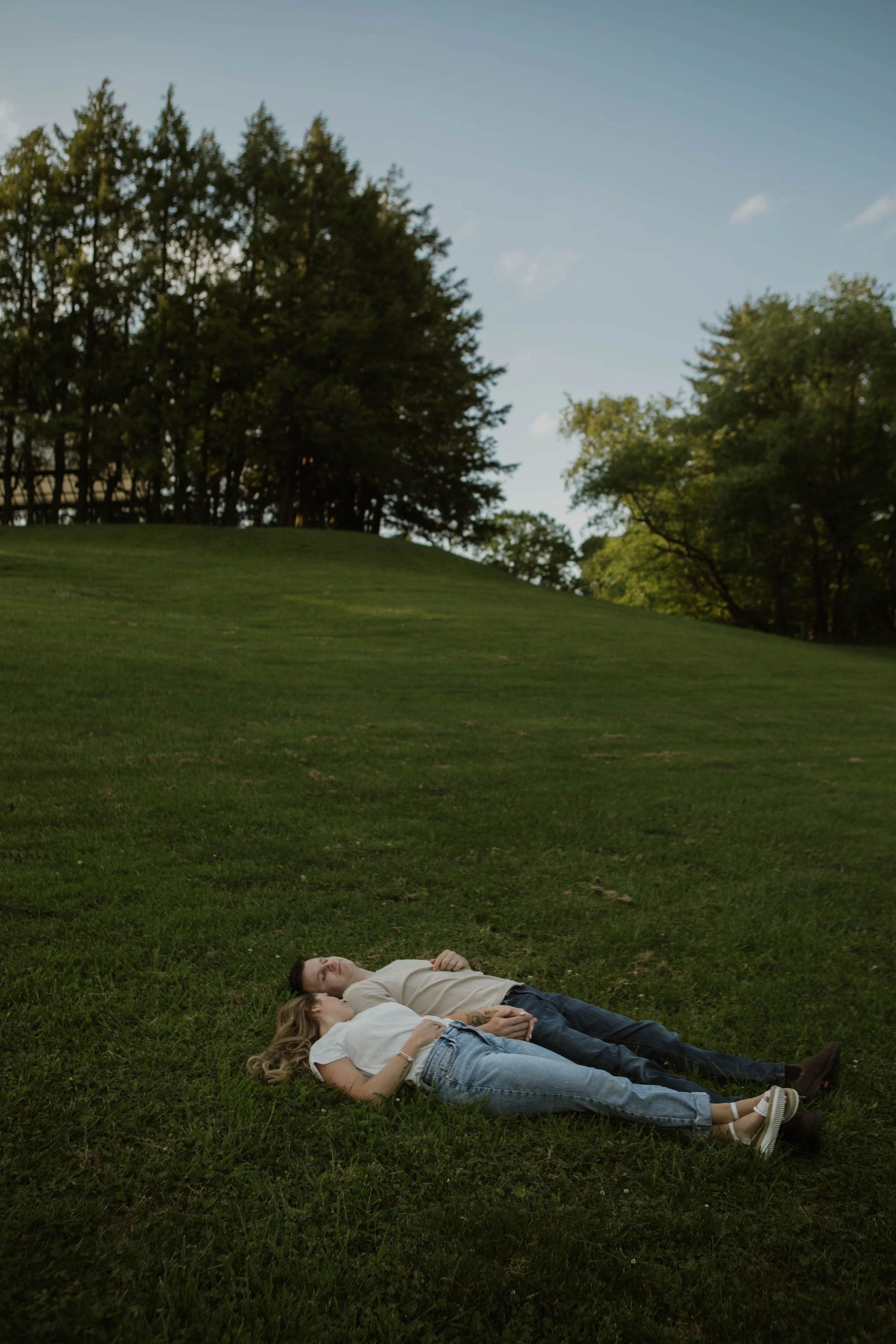 A couple lying on the grass in a park with trees and a blue sky in the background.