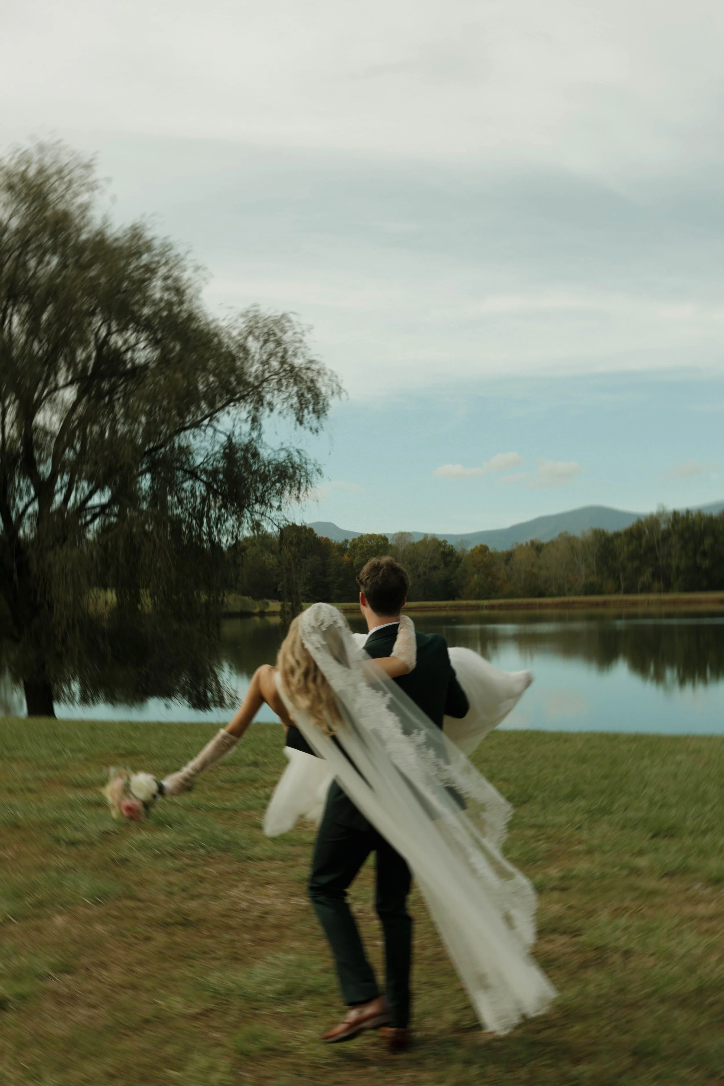 A couple, the man in a dark suit and the woman in a wedding dress with a long veil, walking by a lake, with the man carrying the woman in his arms, holding a bouquet of flowers, during daytime.