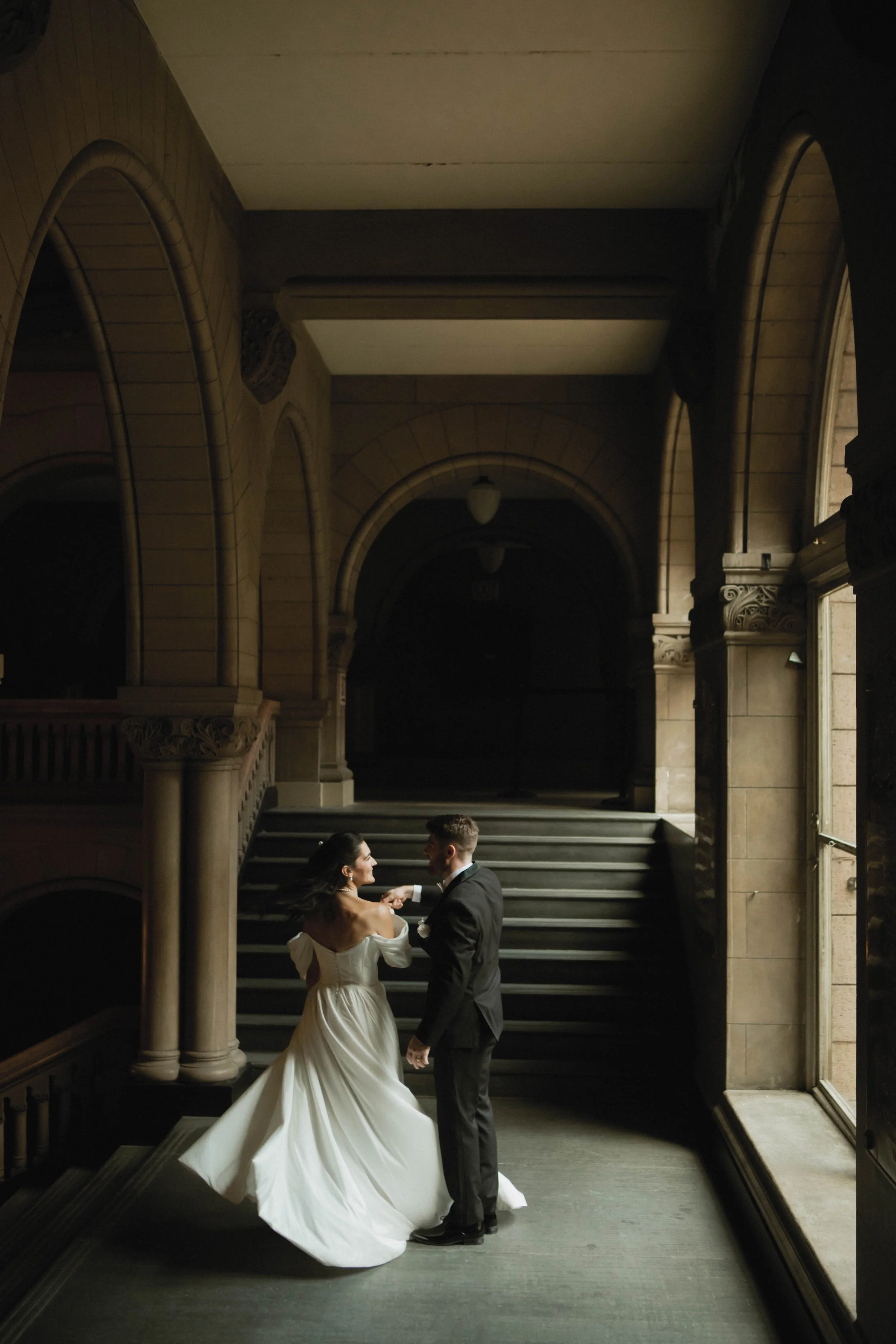 A bride in a white wedding gown and a groom in a black suit dance together in an elegant, historic building with large arched windows and a staircase.