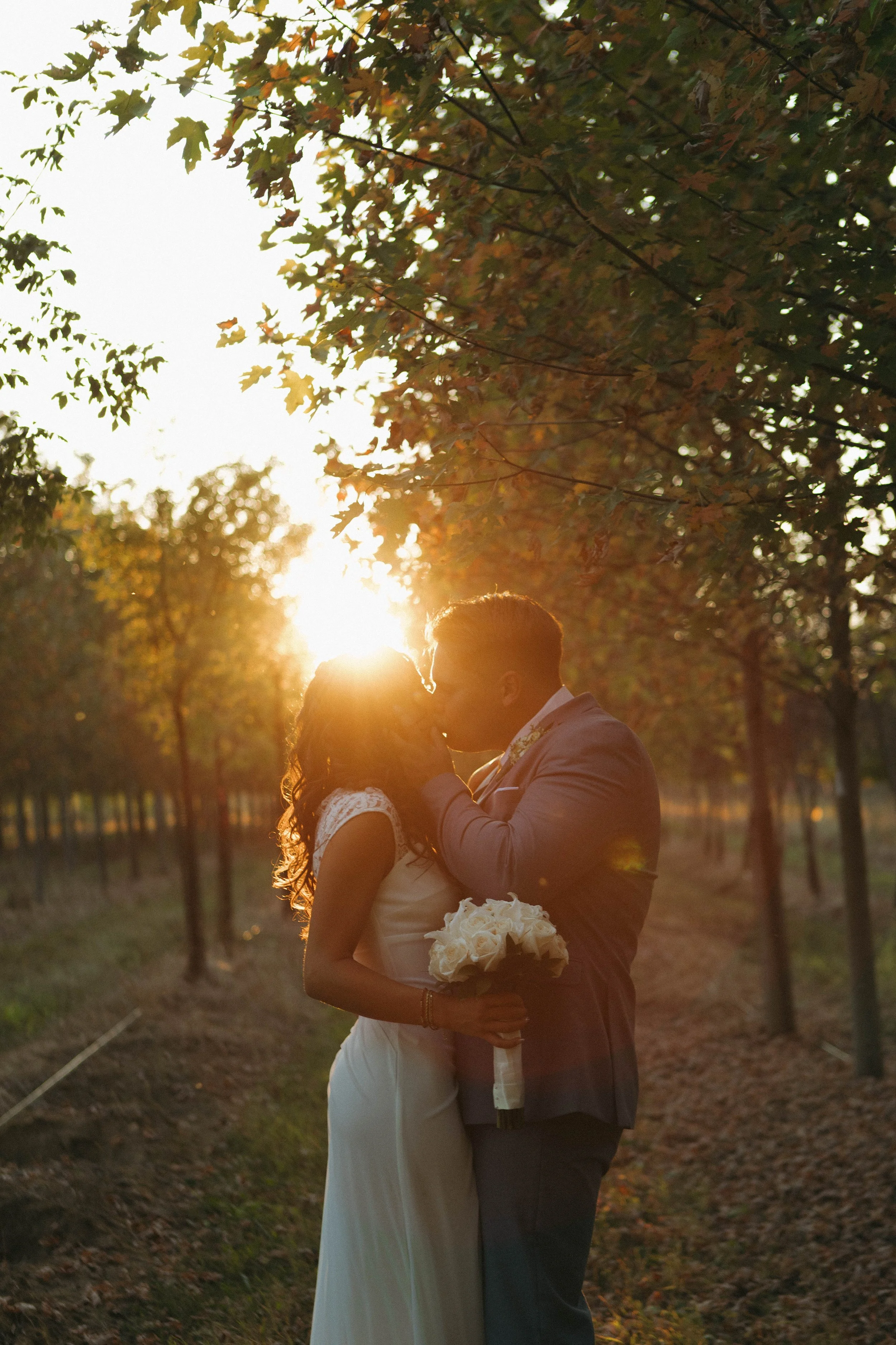 A couple kissing in a tree-lined outdoor setting during sunset, with the woman holding a bouquet of white roses.