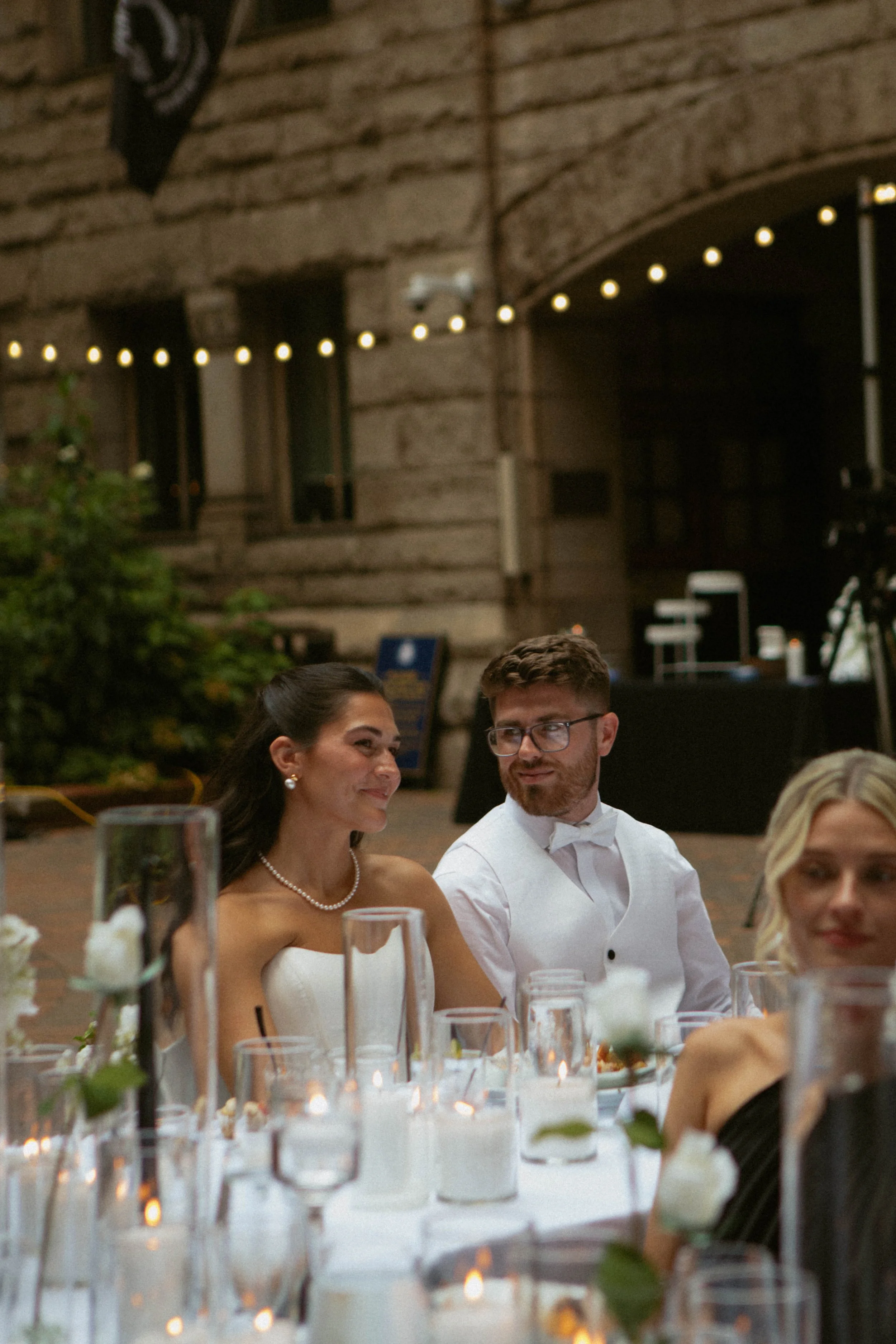 A bride and groom sit at a wedding reception table, smiling and looking at each other, with decorated tables and candles in the foreground and a rustic building with string lights in the background.