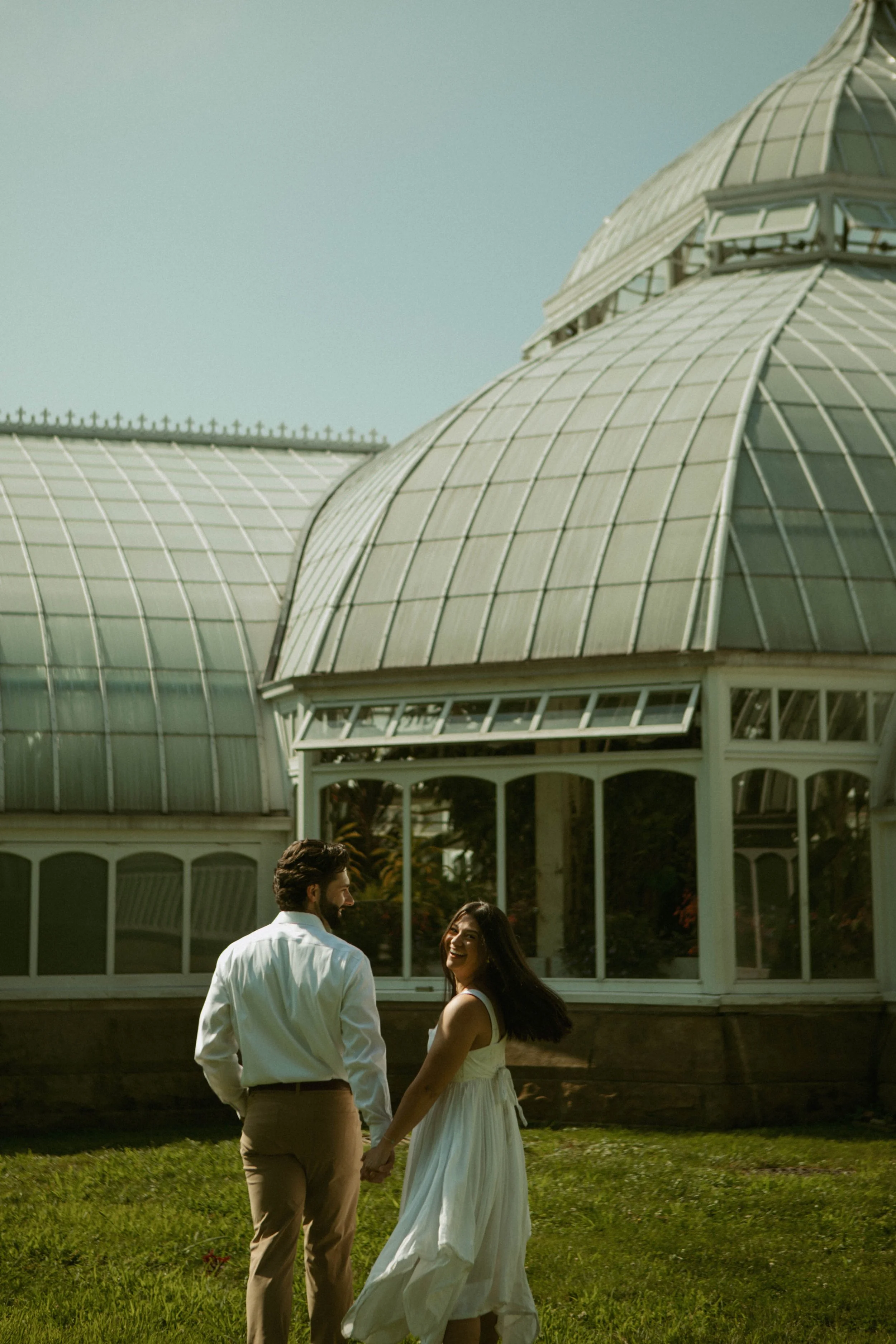 A couple holding hands and smiling outside of a glass greenhouse with domed roofs on a sunny day.