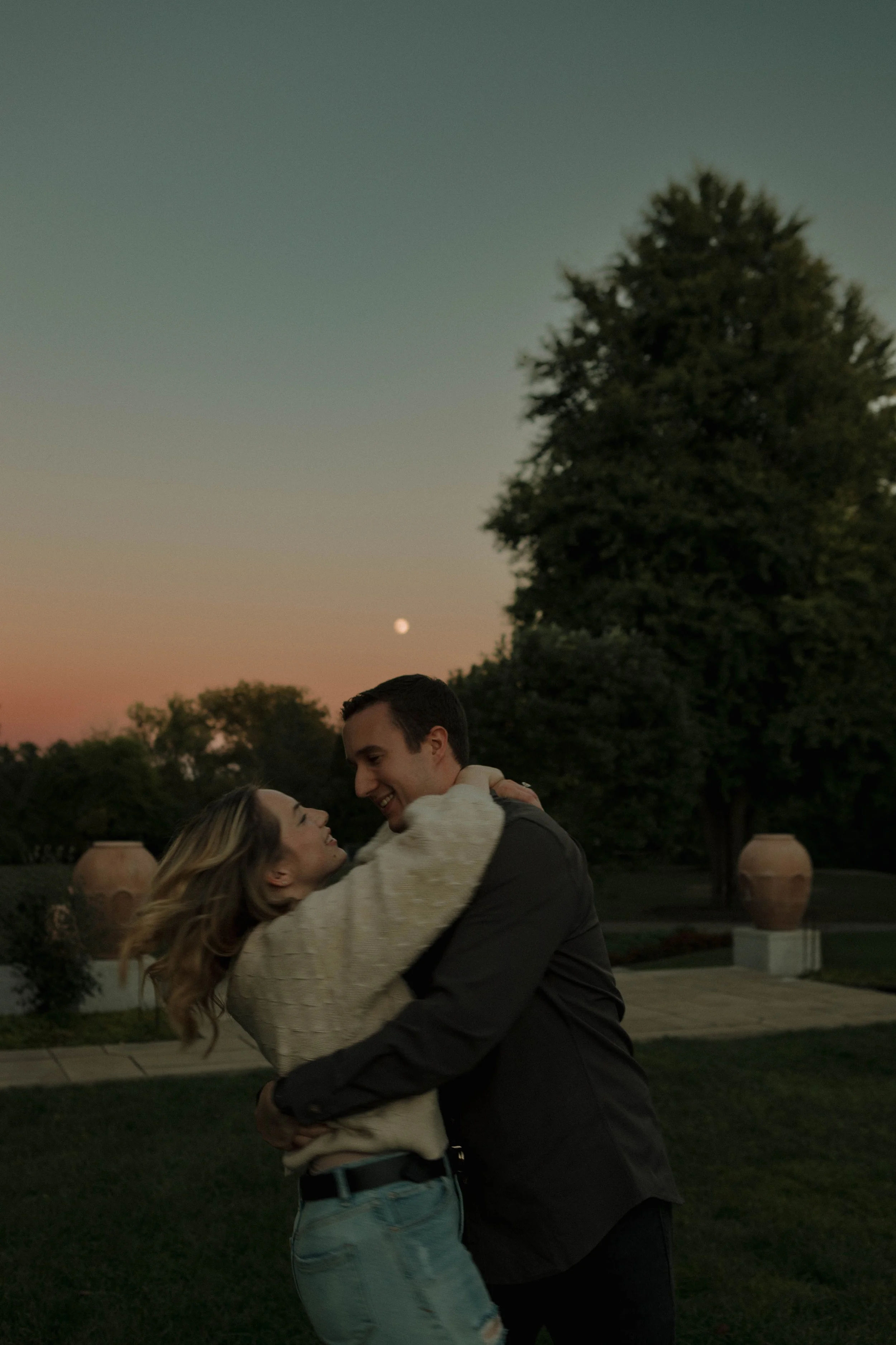 A couple dancing outdoors during sunset, with a tree and the moon in the background, smiling and embracing each other.