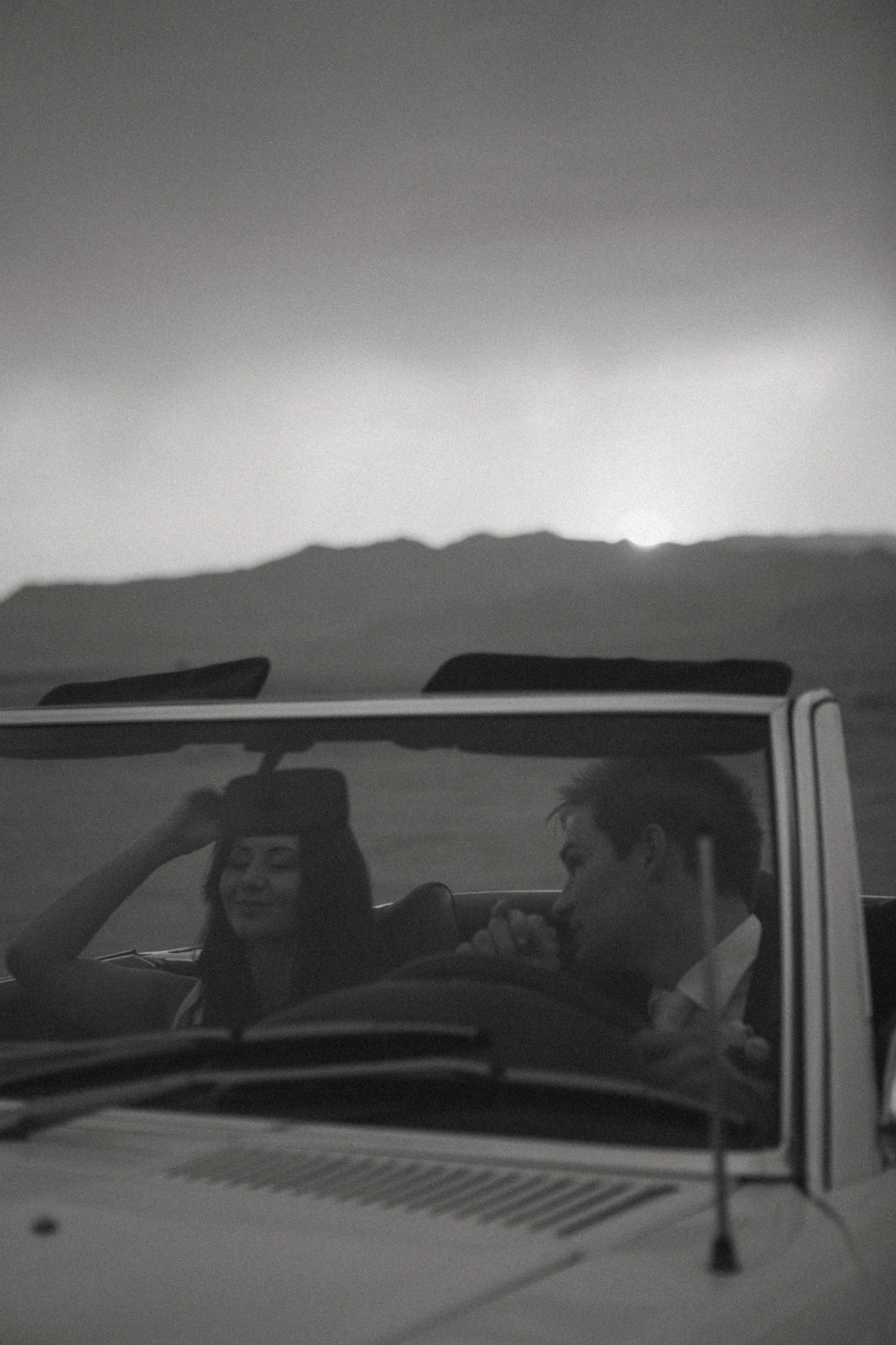 A black and white photo of a young woman and a young man enjoying a drive in a convertible car in a remote, mountainous area during sunset.