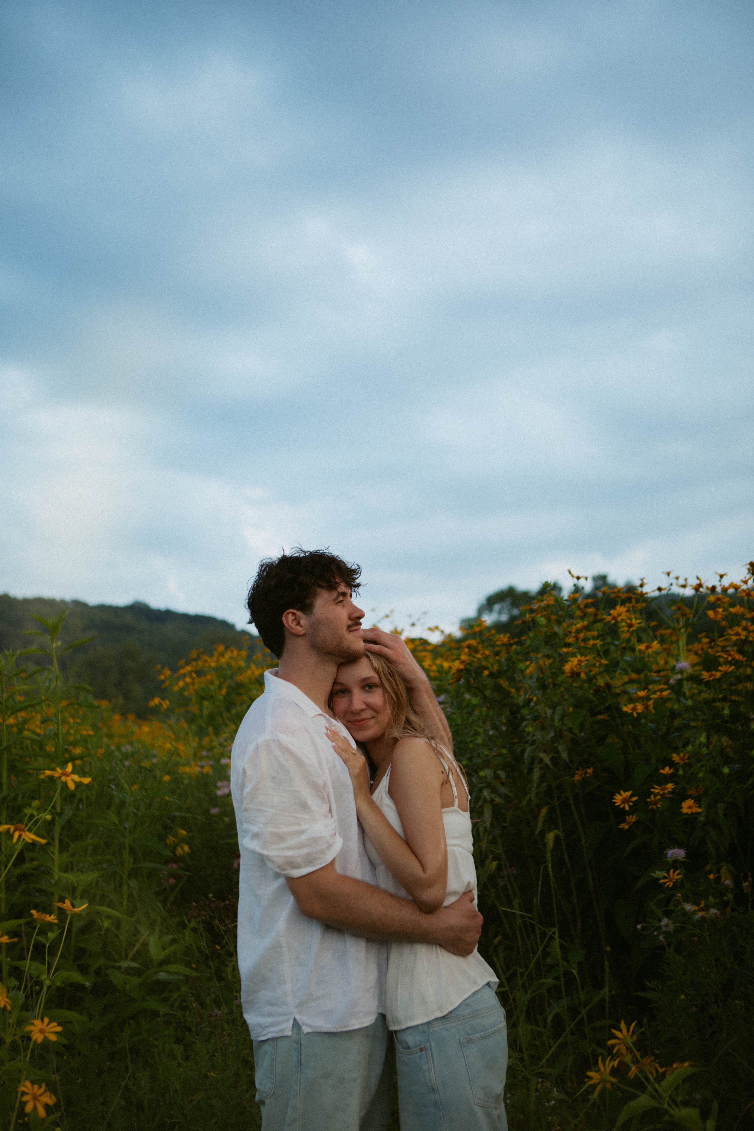 A young couple in white clothing embracing outdoors among yellow flowers with hills and cloudy sky in the background.