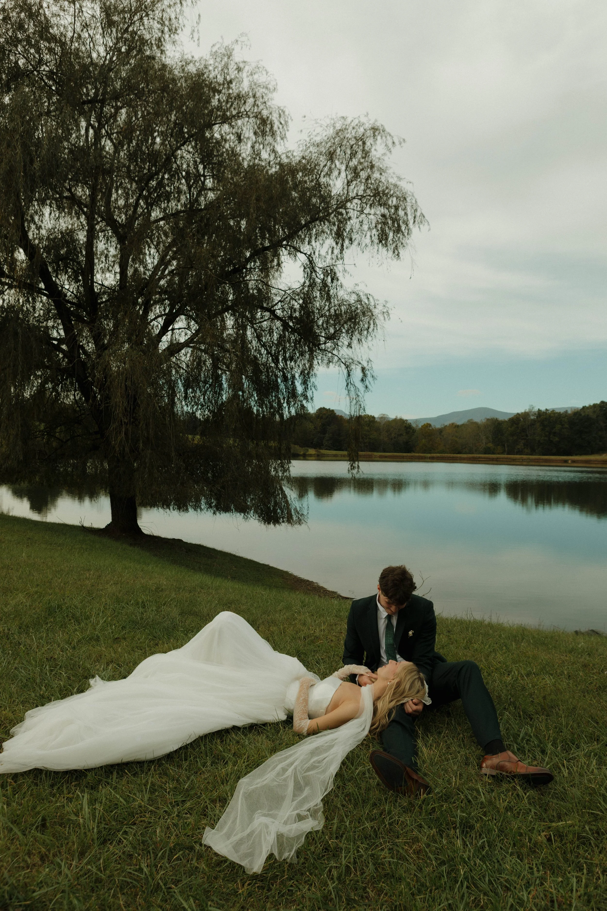 A bride and groom sitting and lying on the grass near a lake, with the bride lying down with her eyes closed and the groom looking at her, under a large tree on a cloudy day.