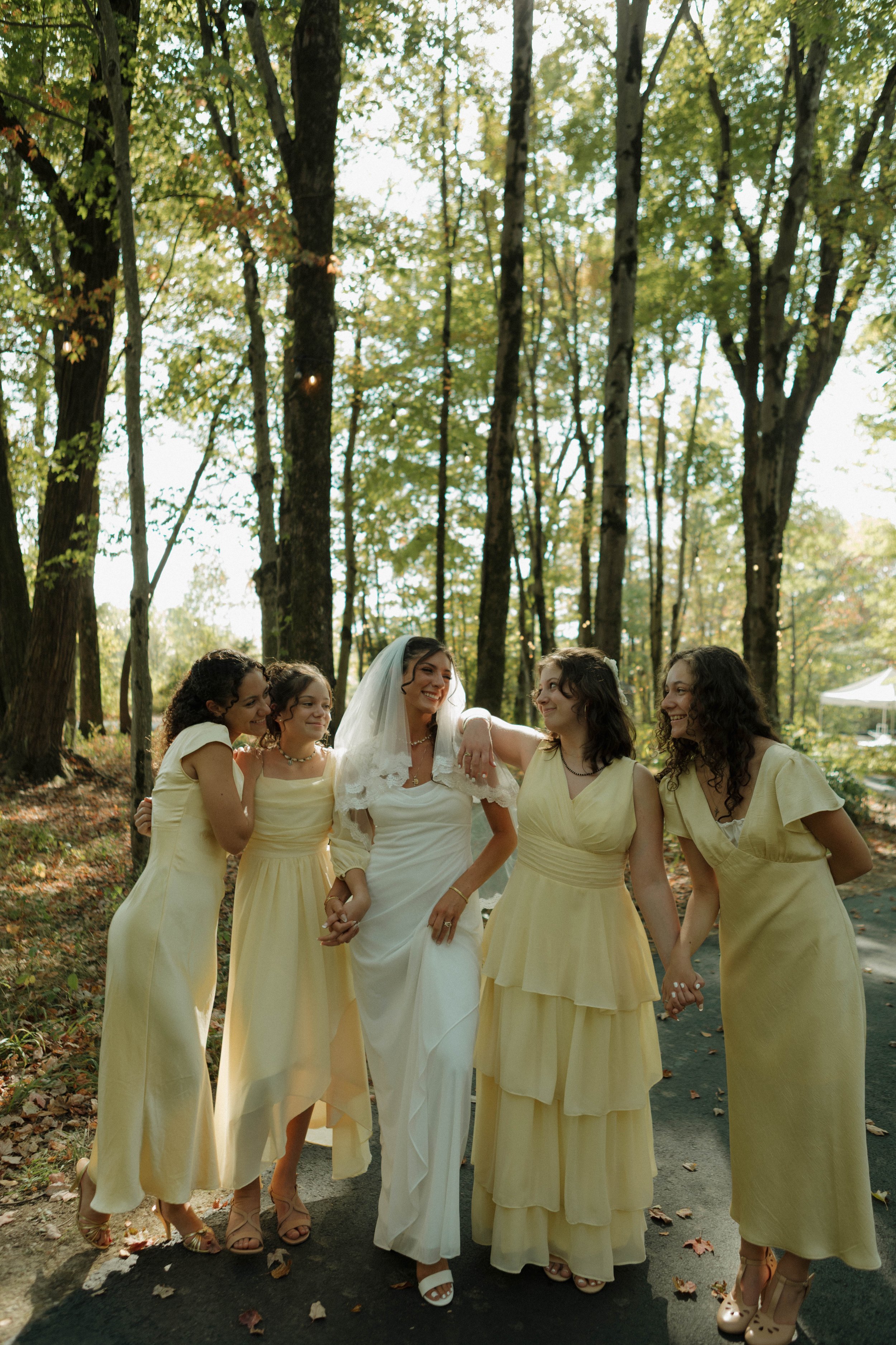 A bride and her bridesmaids walking through a wooded area, holding hands and smiling, during a wedding celebration.