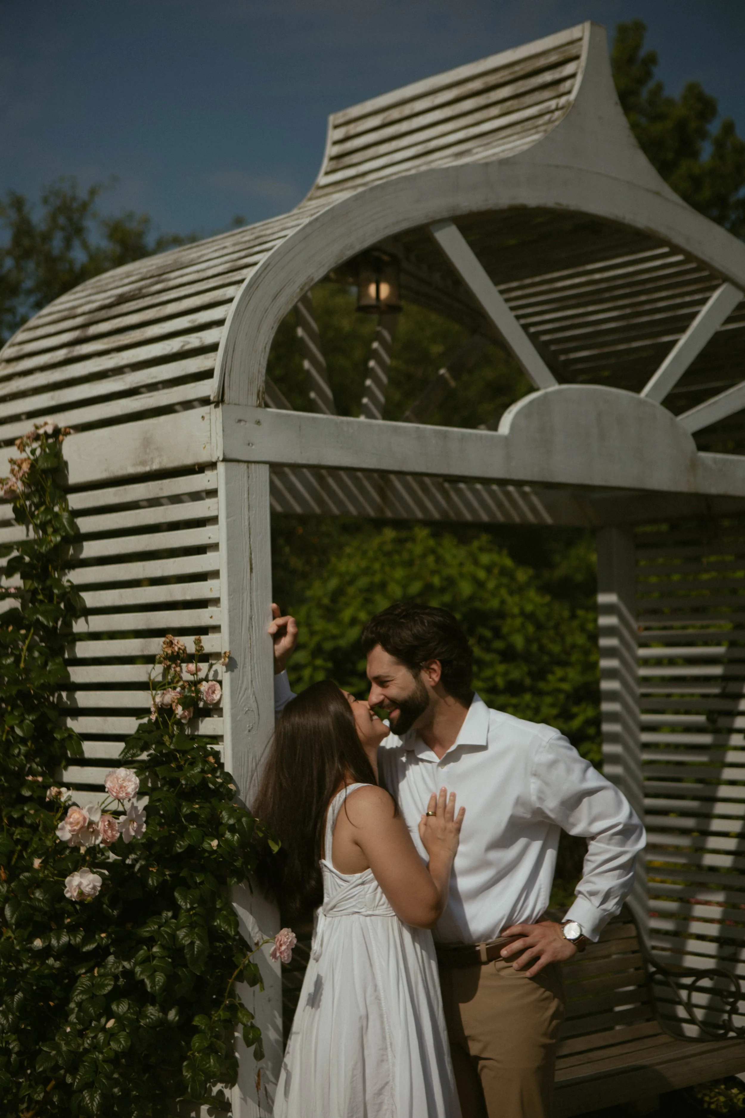A couple is kissing under a white wooden gazebo with pink flowers growing nearby, during daytime.
