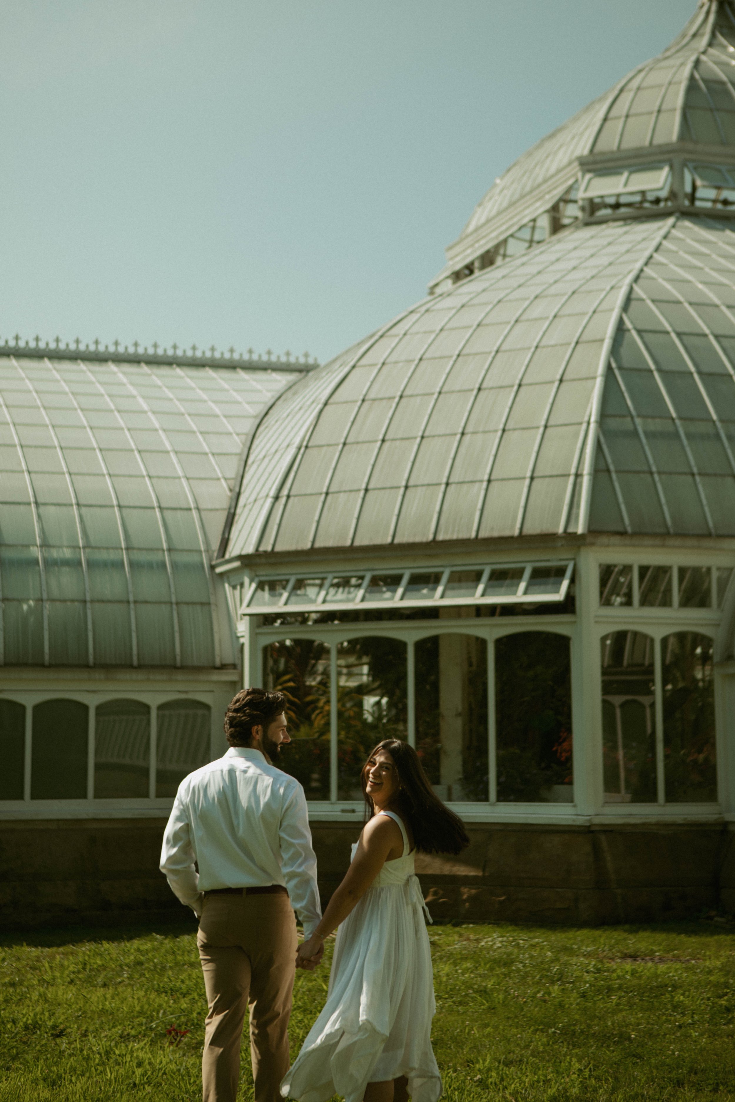 A couple holds hands and walks in front of a historic glass greenhouse, smiling and looking at each other on a sunny day.