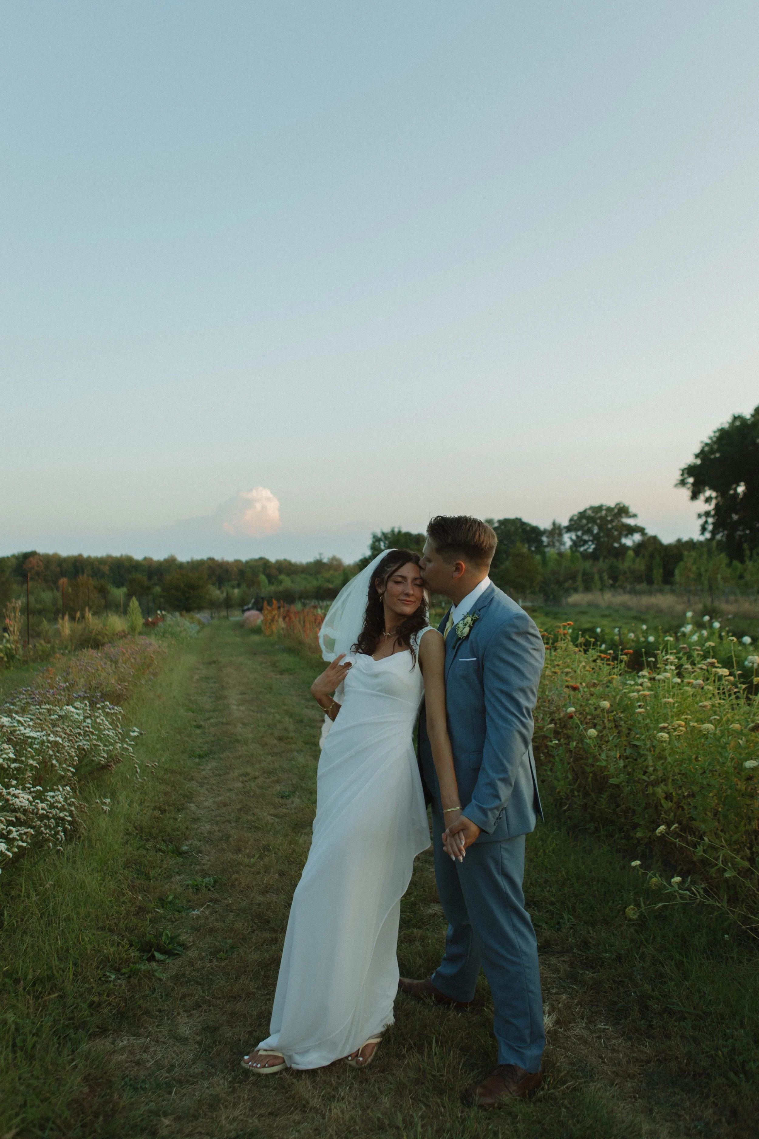 A bride and groom holding hands in a field at sunset, with the groom kissing the bride on the forehead.