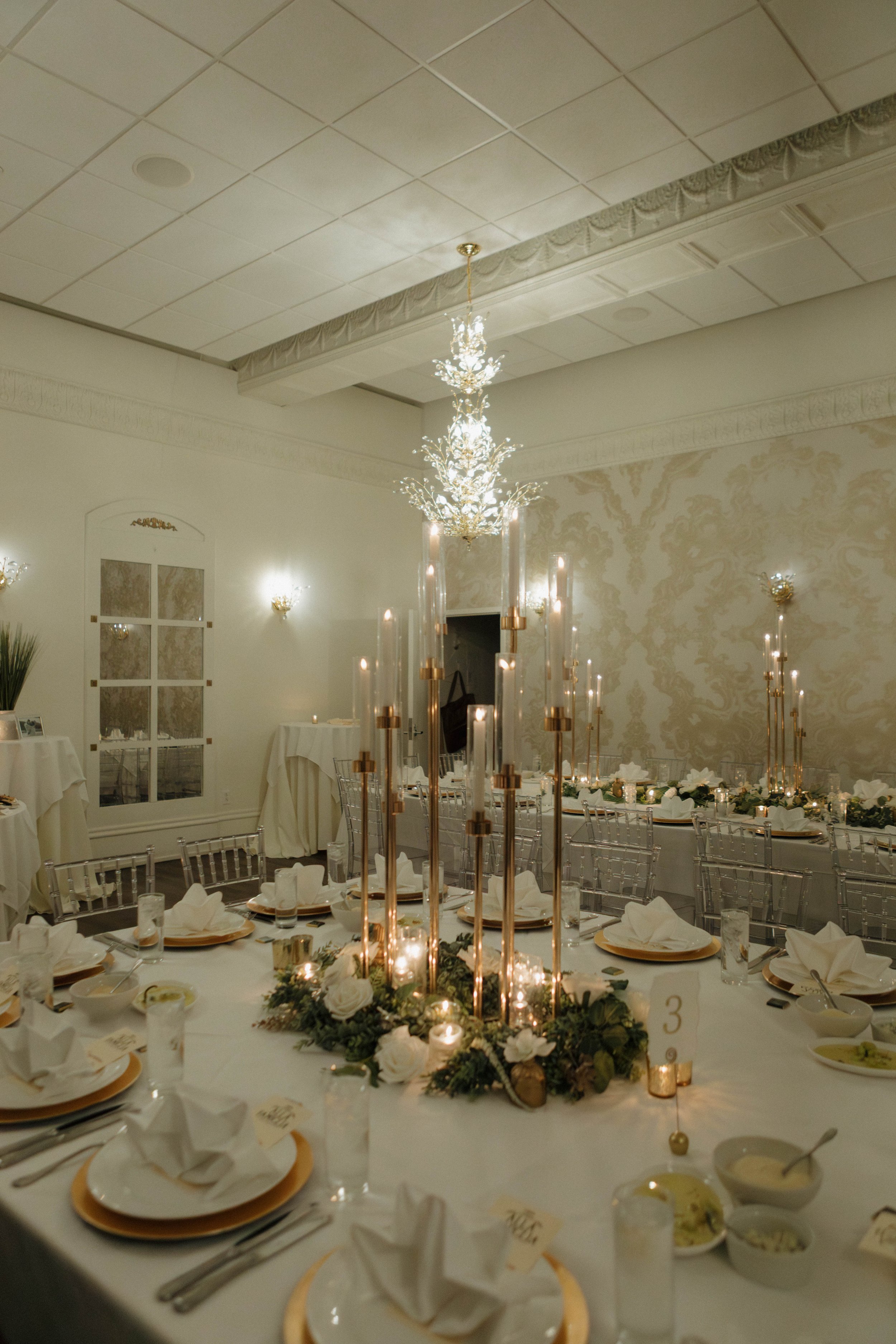 Elegant banquet table setting with tall gold candle holders, white floral arrangements, and neatly folded white napkins at each place setting, in a decorated room with chandeliers and soft lighting.