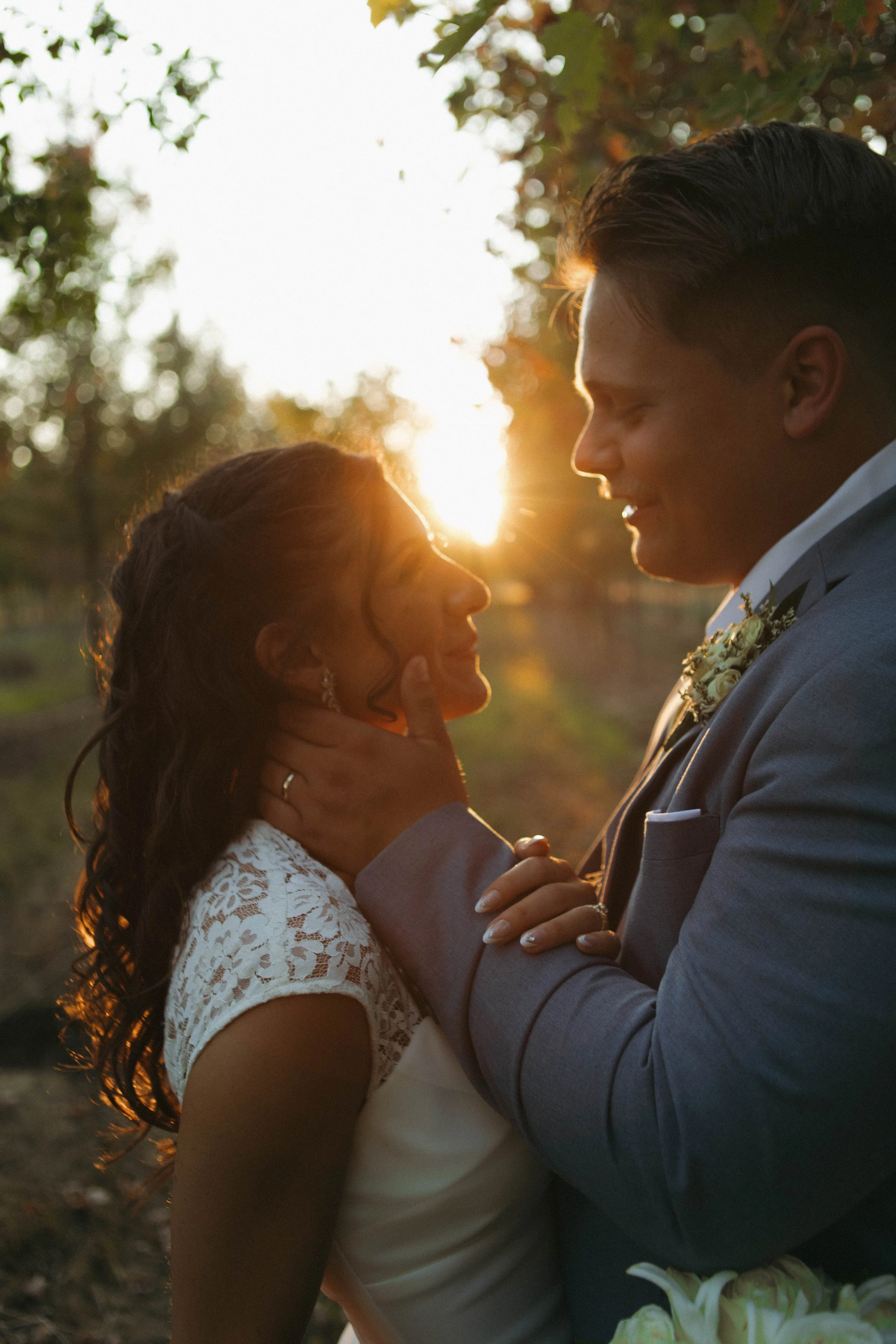 A couple dressed in wedding attire standing close together outdoors during sunset, with the sun shining between them, creating a romantic silhouette.