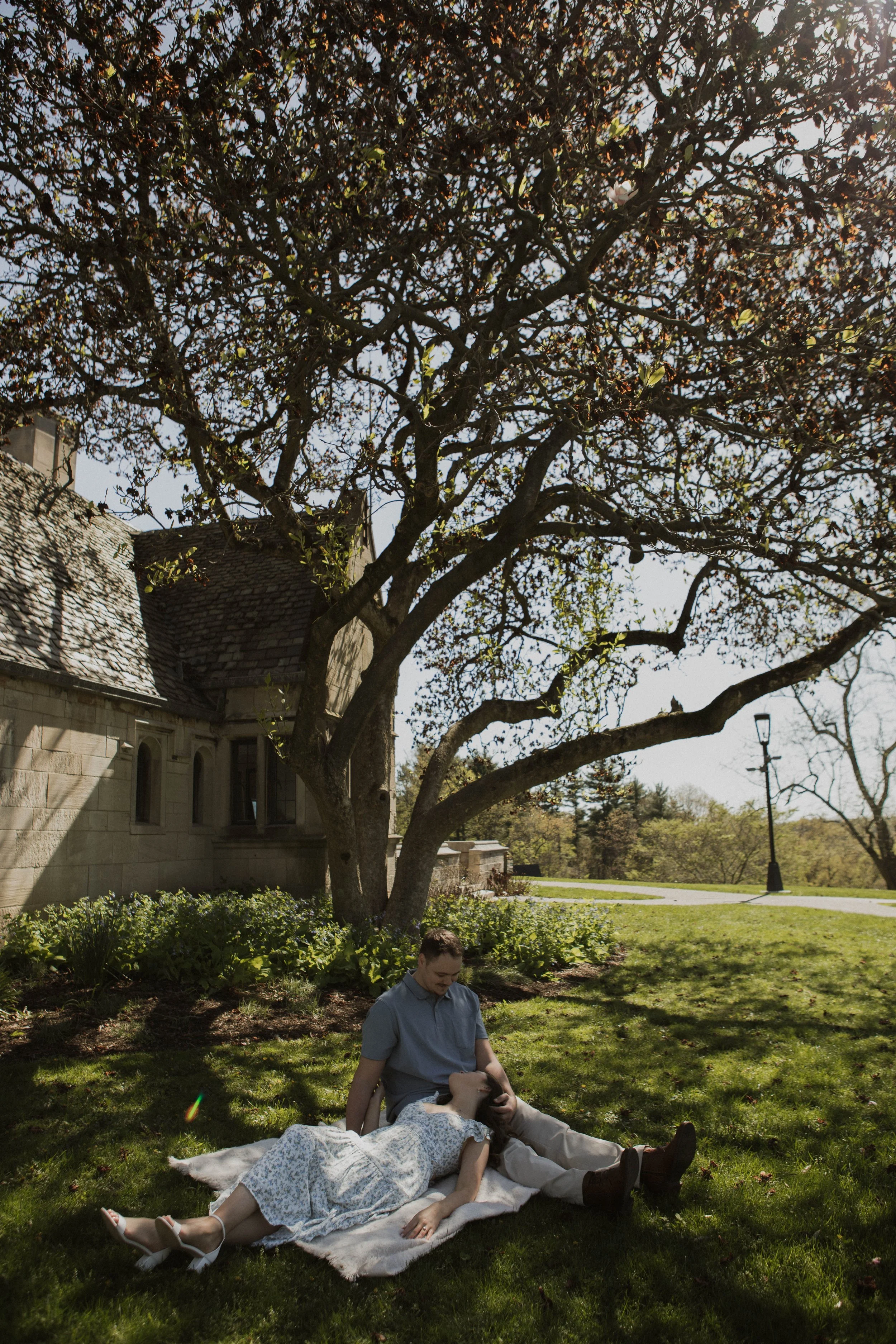 A couple sitting on a blanket under a tree on a sunny day, with a building and a lamppost in the background.