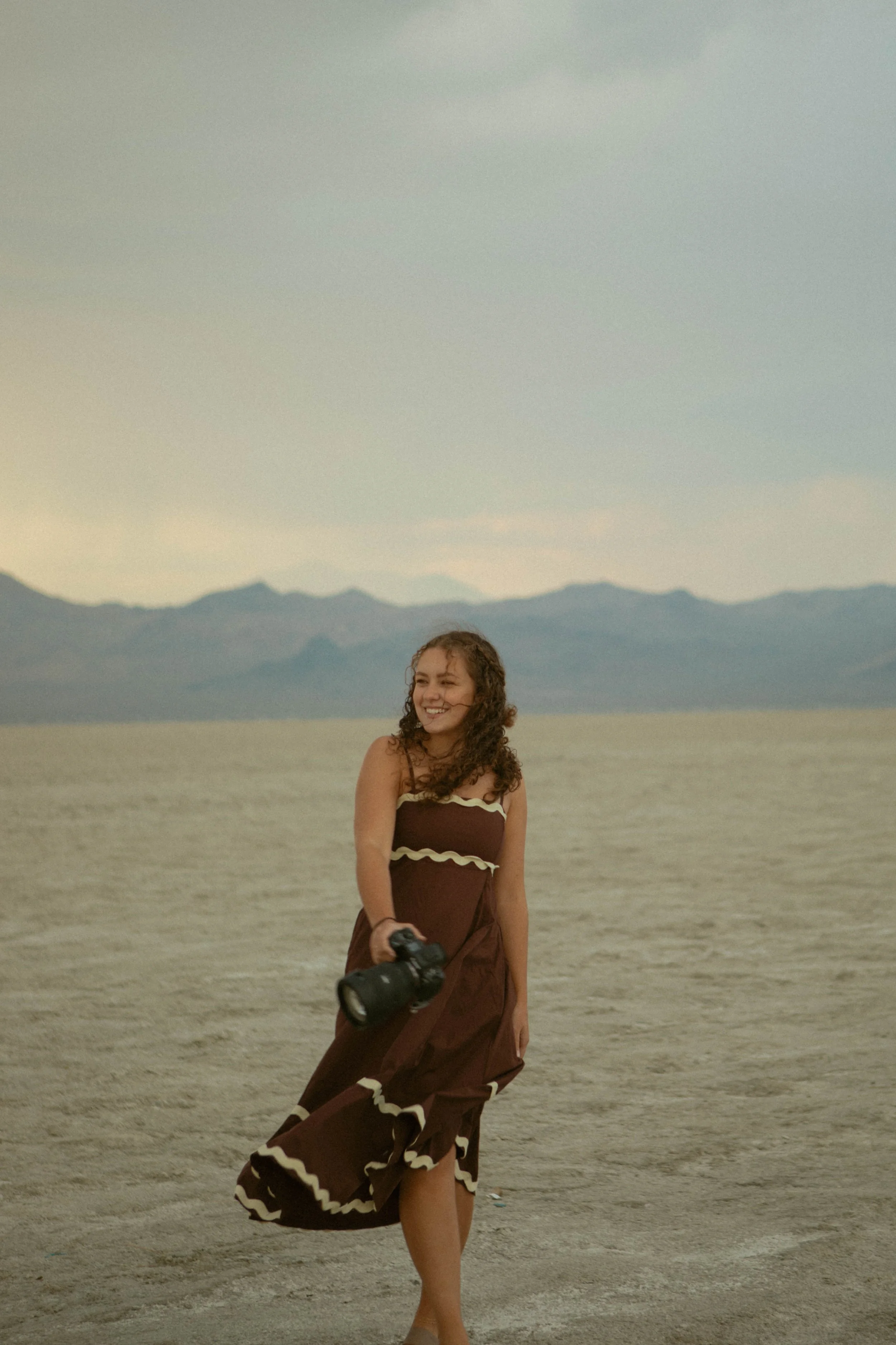 A woman in a brown dress with cream-colored trim holding a camera, standing in a dry, desert-like landscape with mountains in the background.