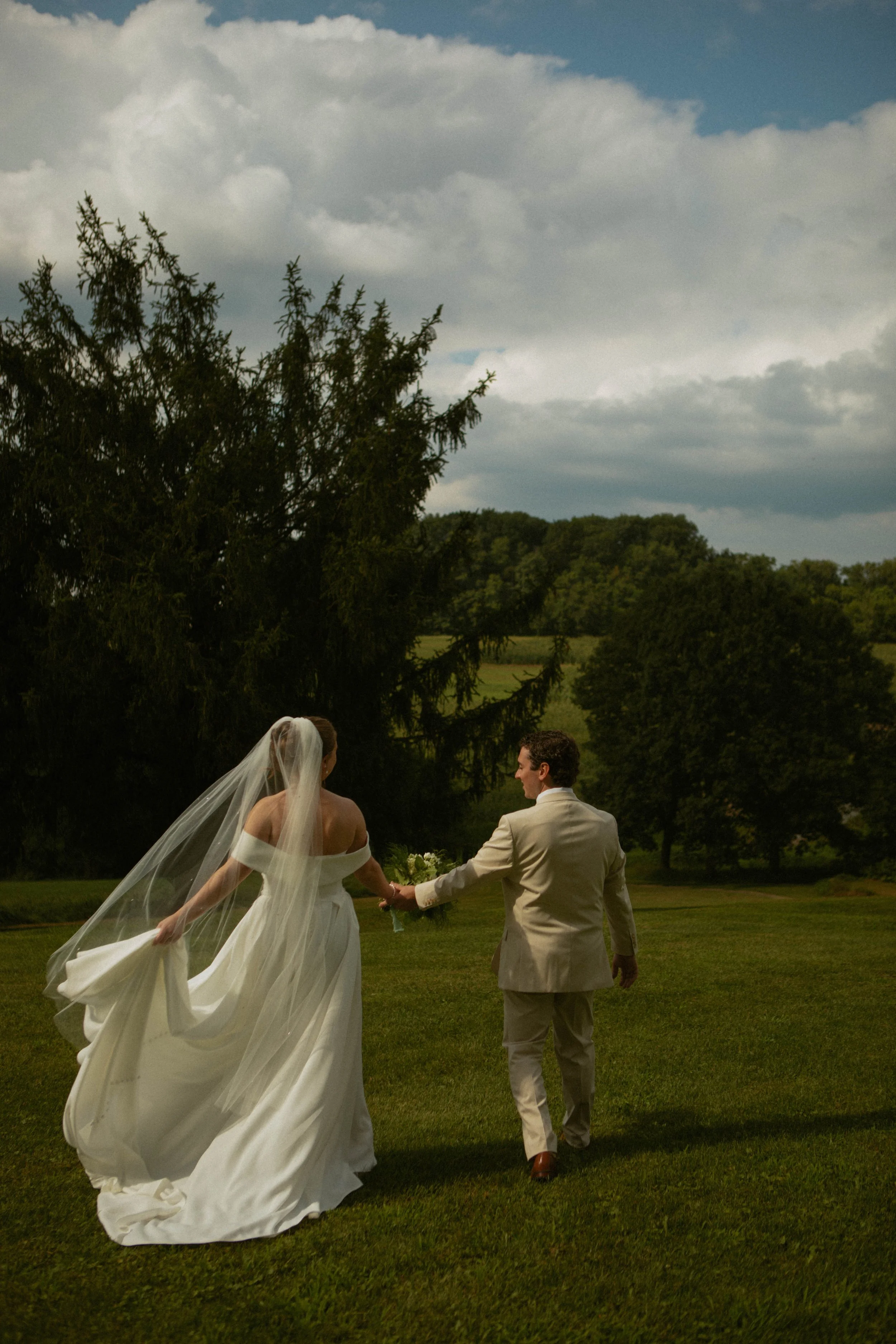A bride and groom holding hands walking on a grassy field during daytime, with a large tree and cloudy sky in the background.