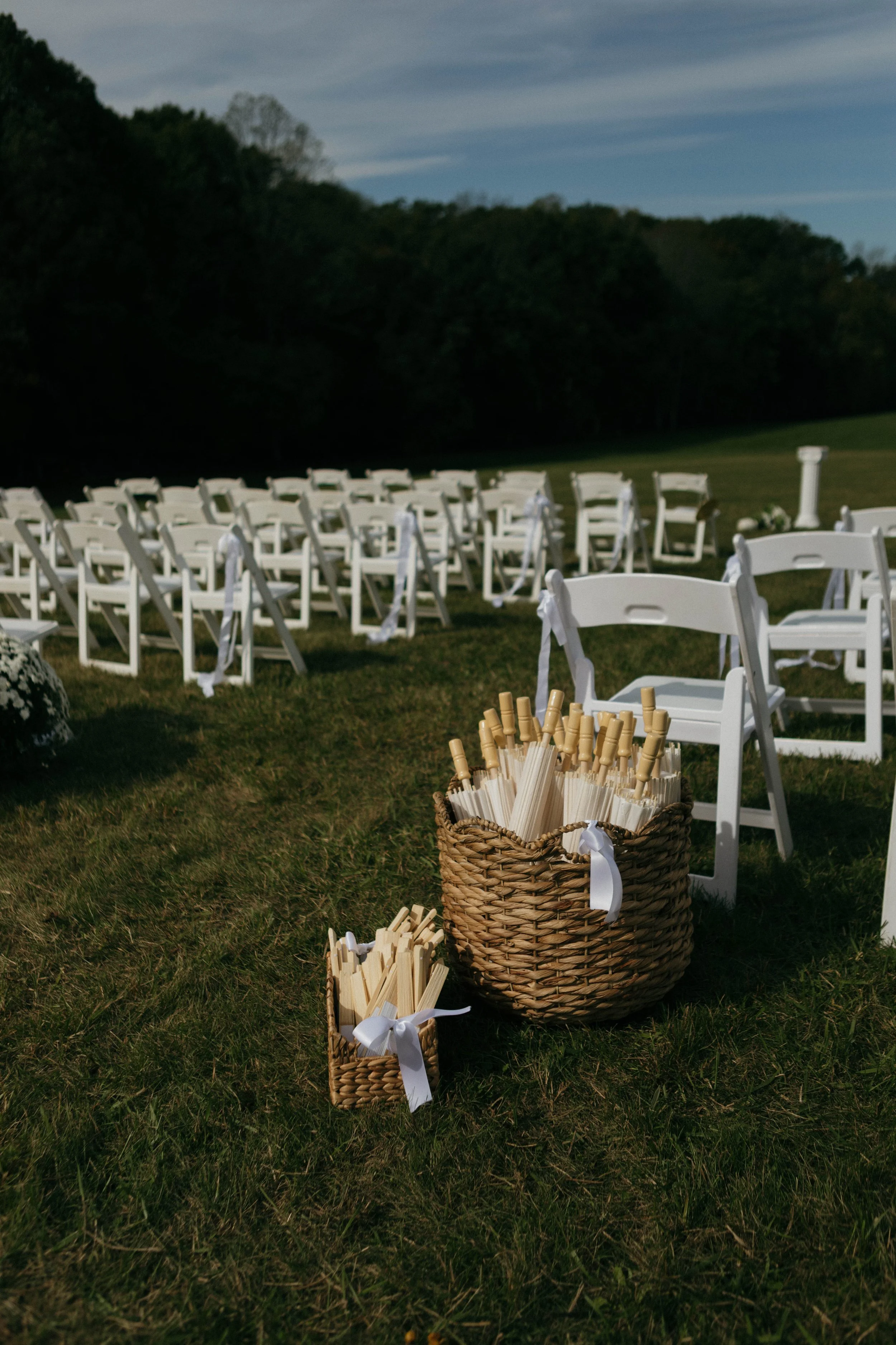 White chairs decorated with white ribbons are arranged outdoors for a wedding ceremony on a grassy field, with baskets of wooden sticks and paper fans in the foreground.