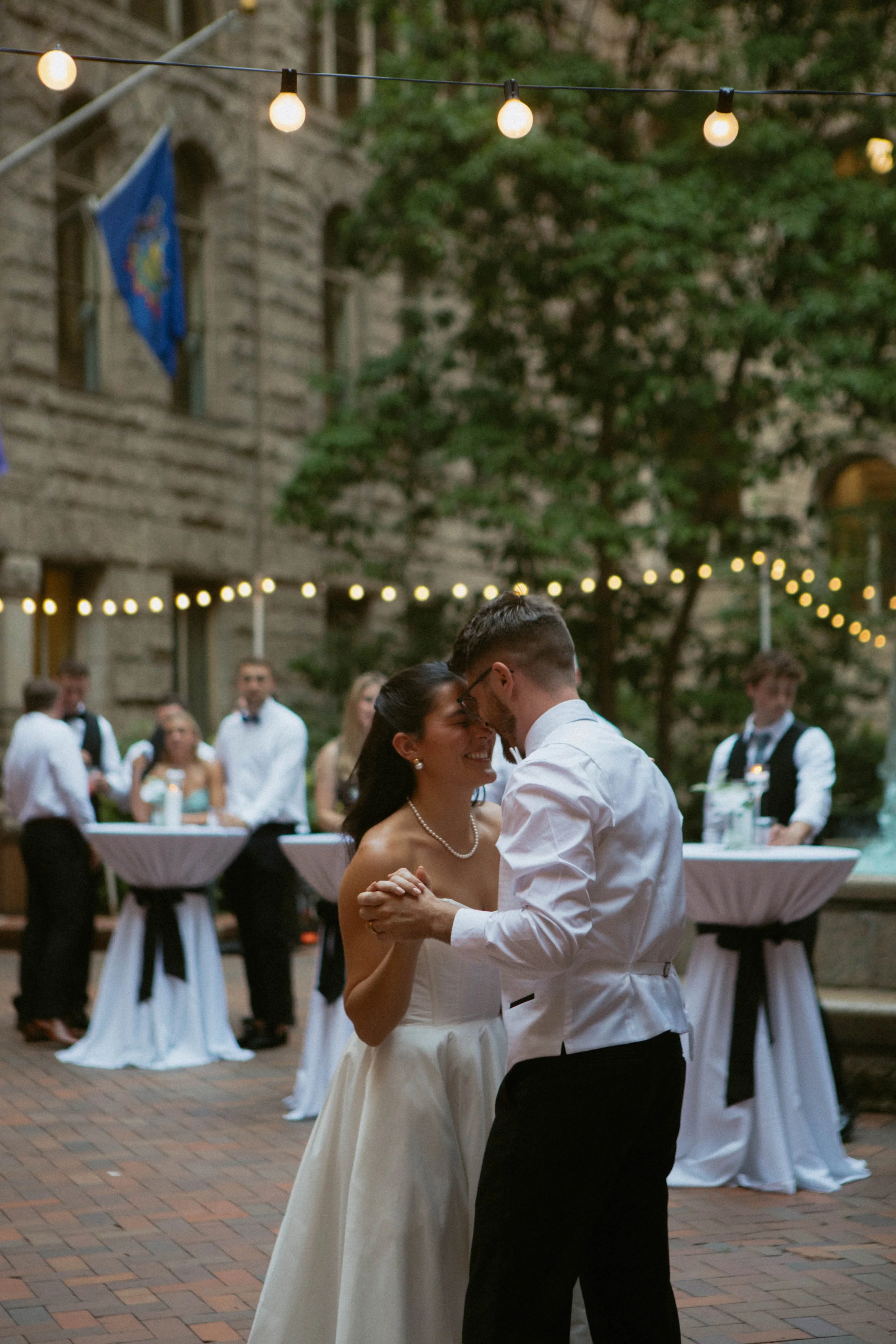 A couple dancing close together during a wedding reception outdoors, with friends in the background, string lights overhead, and a historic stone building behind them.