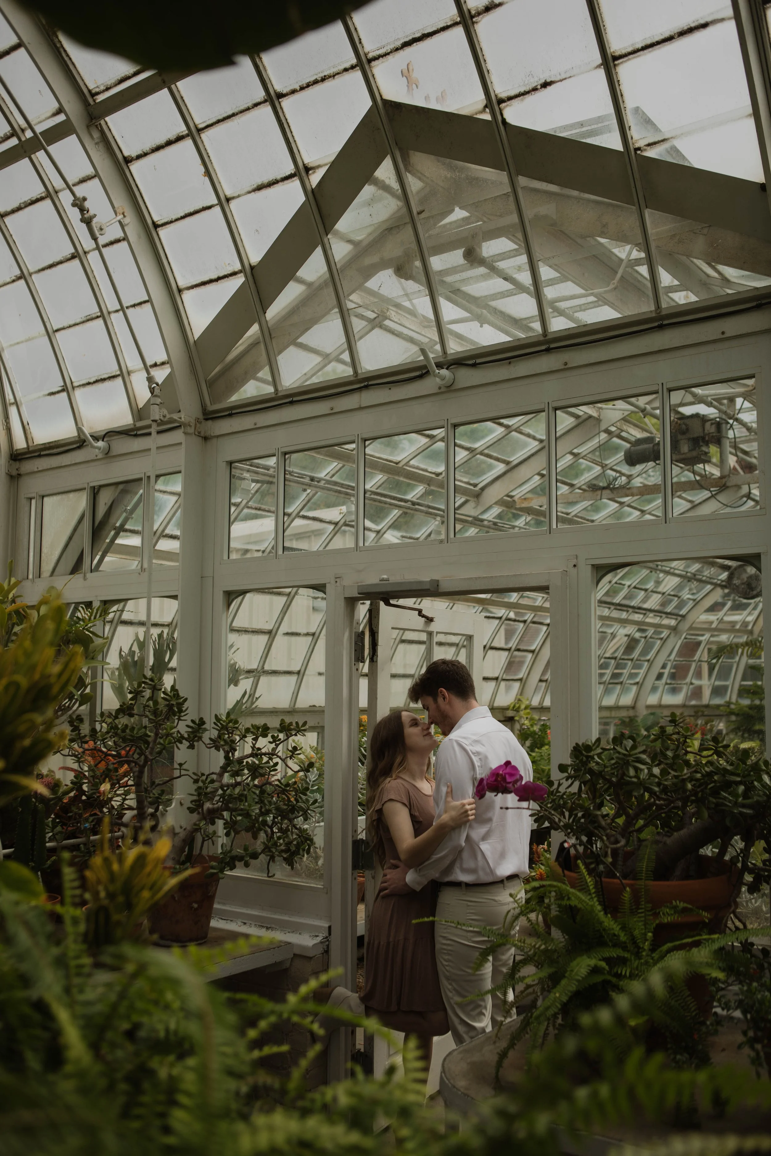 A couple sharing an intimate moment inside a greenhouse surrounded by various plants and flowers.