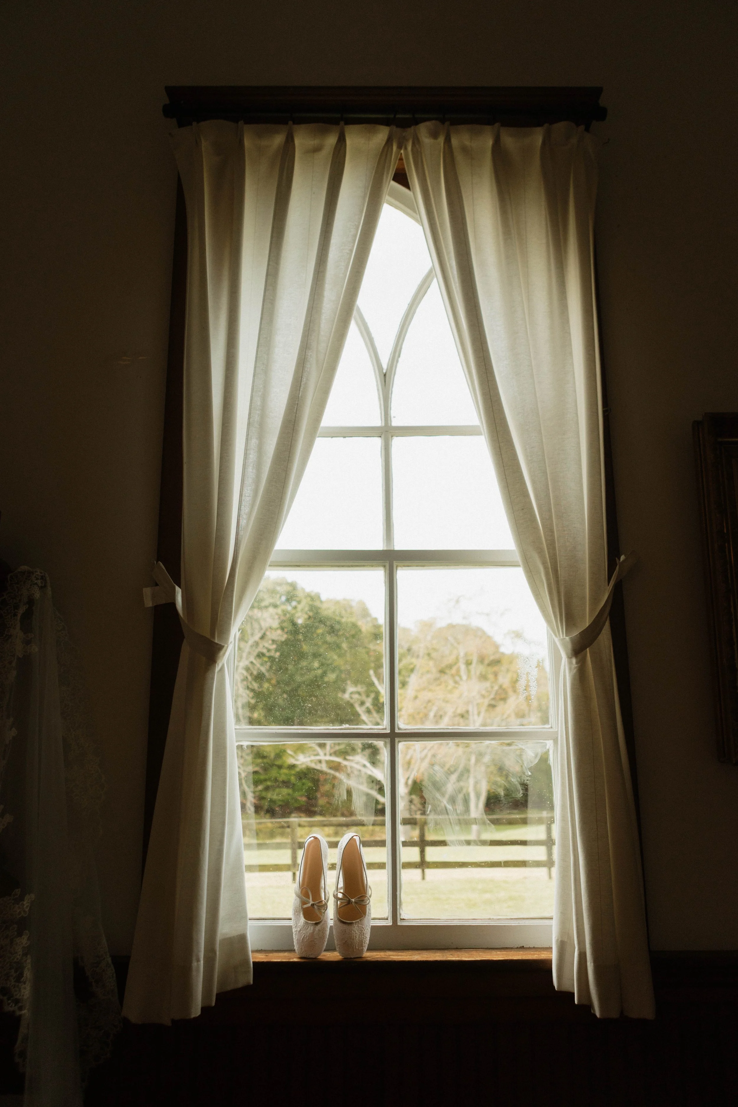 A pair of white wedding shoes placed on a window sill behind a window with white curtains, showing an outdoor landscape with trees and a fence.