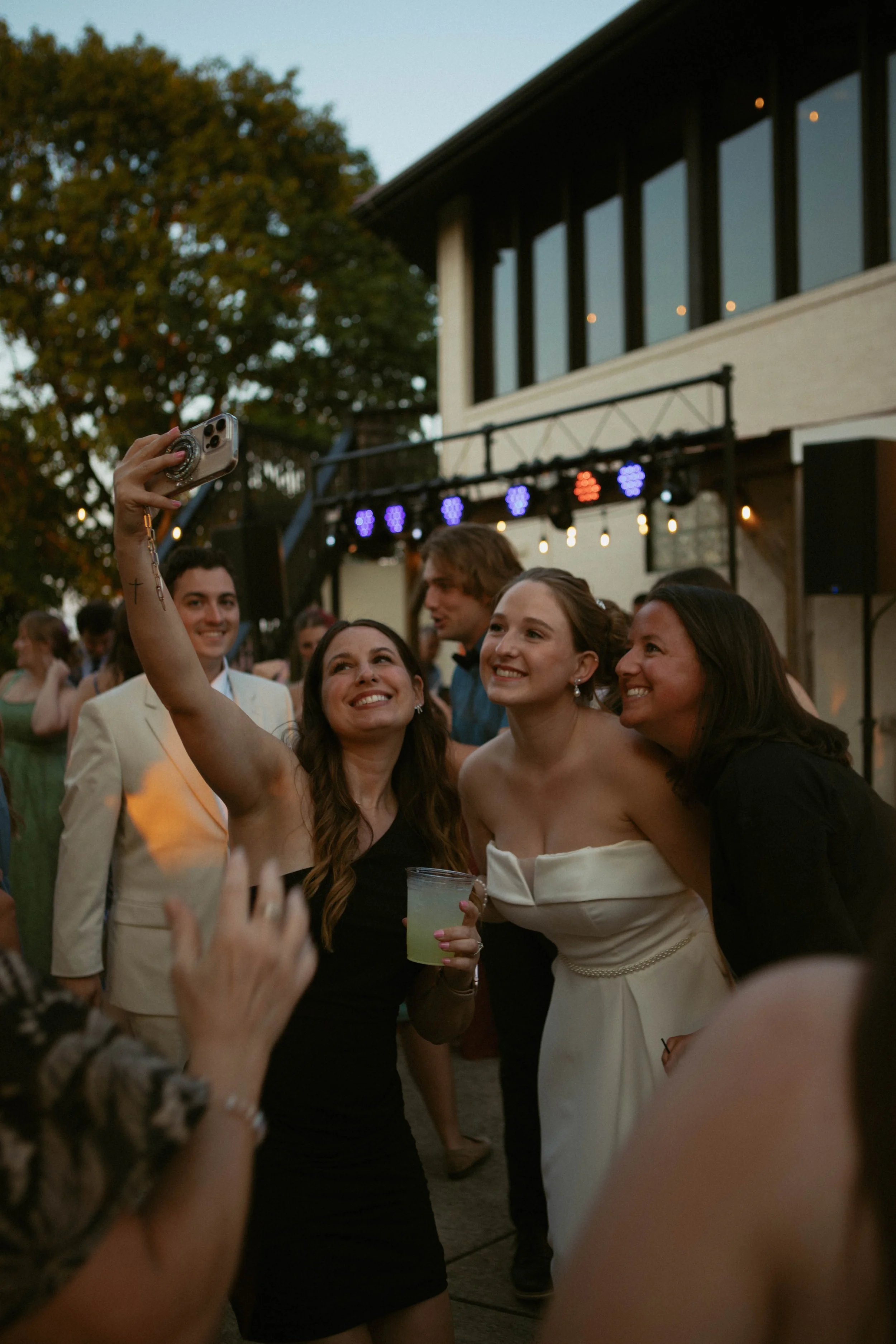 People at an outdoor party taking a selfie together, with a woman in a white dress, smiling and holding a drink, surrounded by friends.