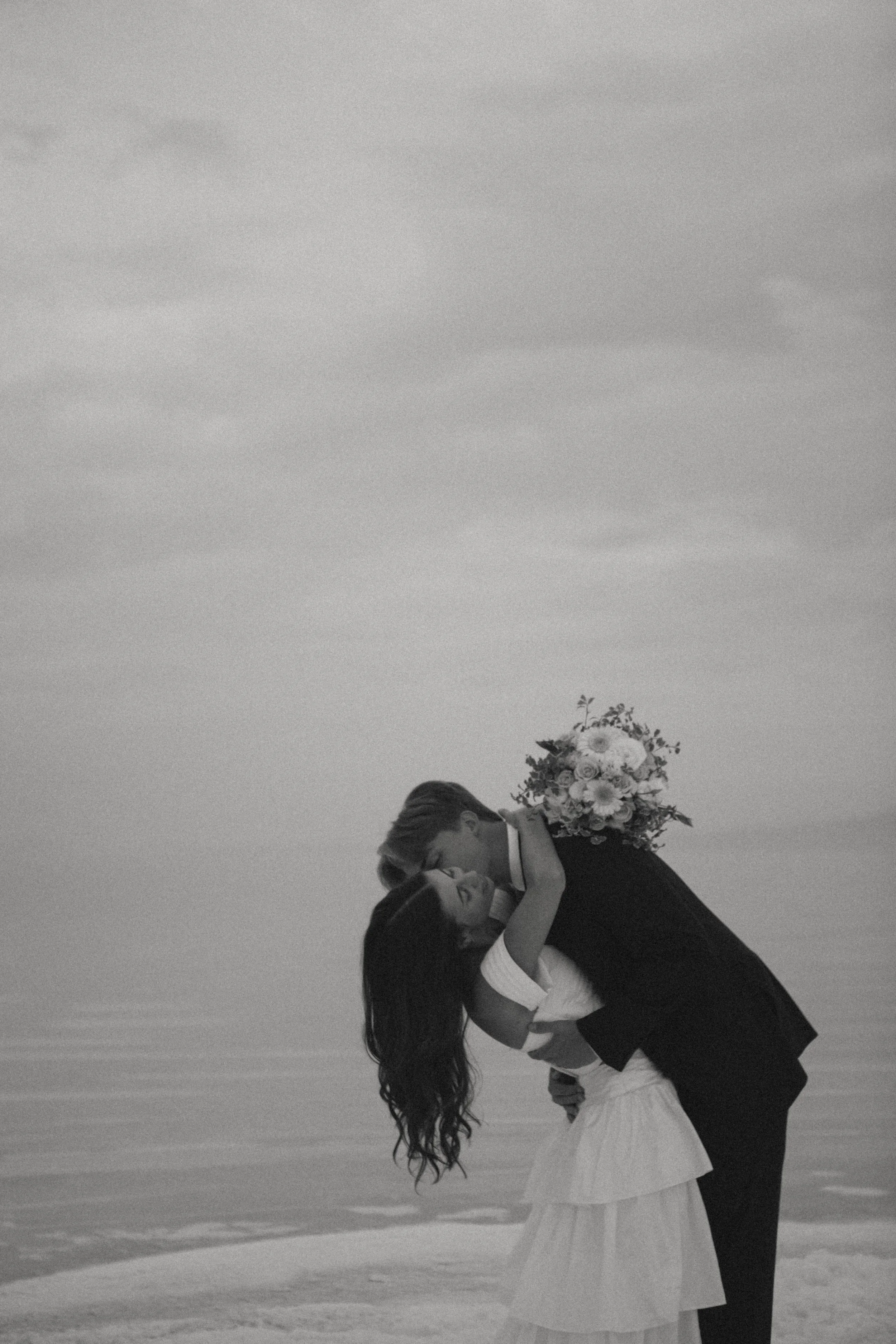 A black and white photo of a couple kissing on the beach, with one person dipping the other. The person being dipped holds a bouquet of flowers, and the background shows the ocean and cloudy sky.