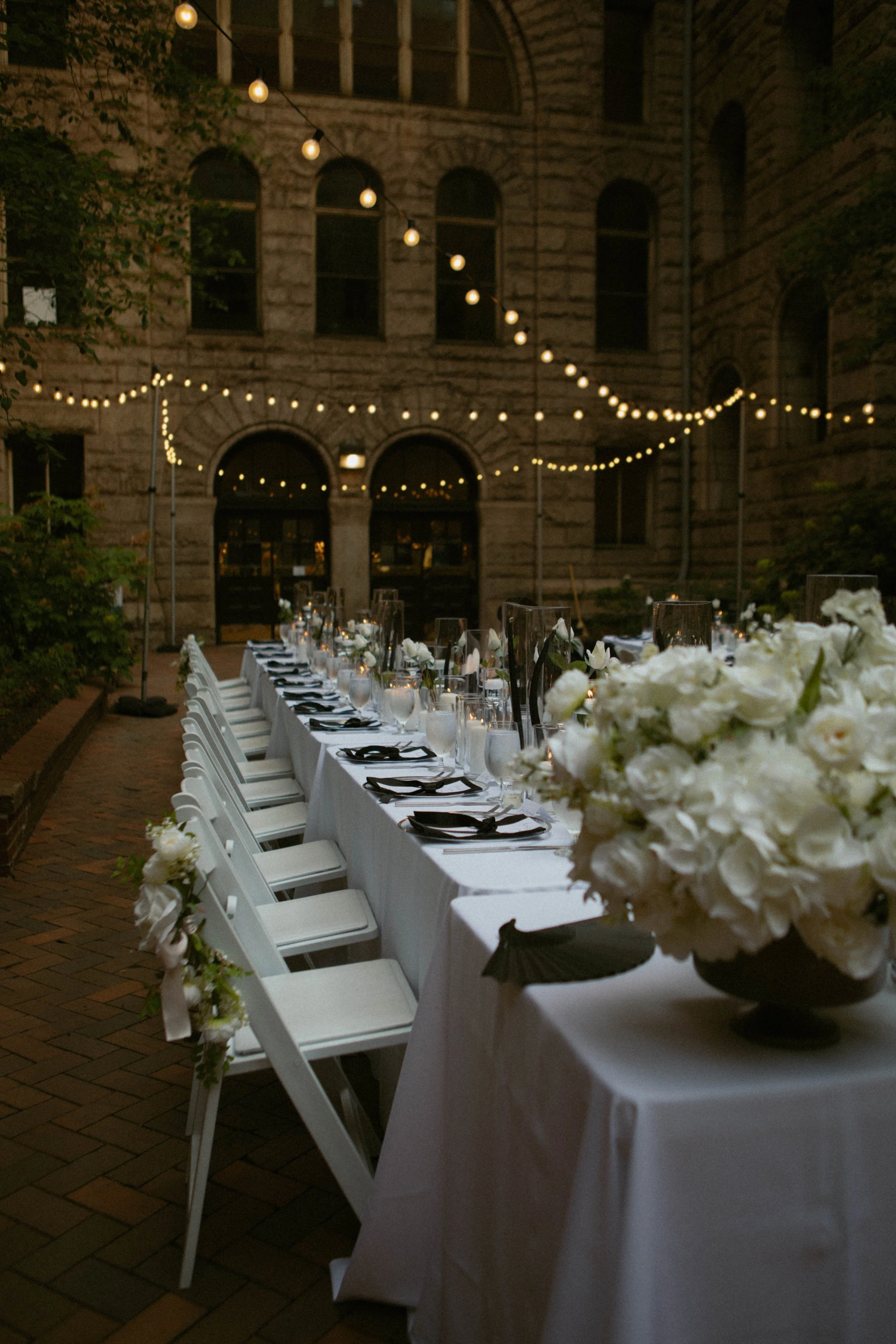 Outdoor dinner table decorated with white floral arrangements, black napkins, and glassware, set in a courtyard with string lights hanging overhead during evening.